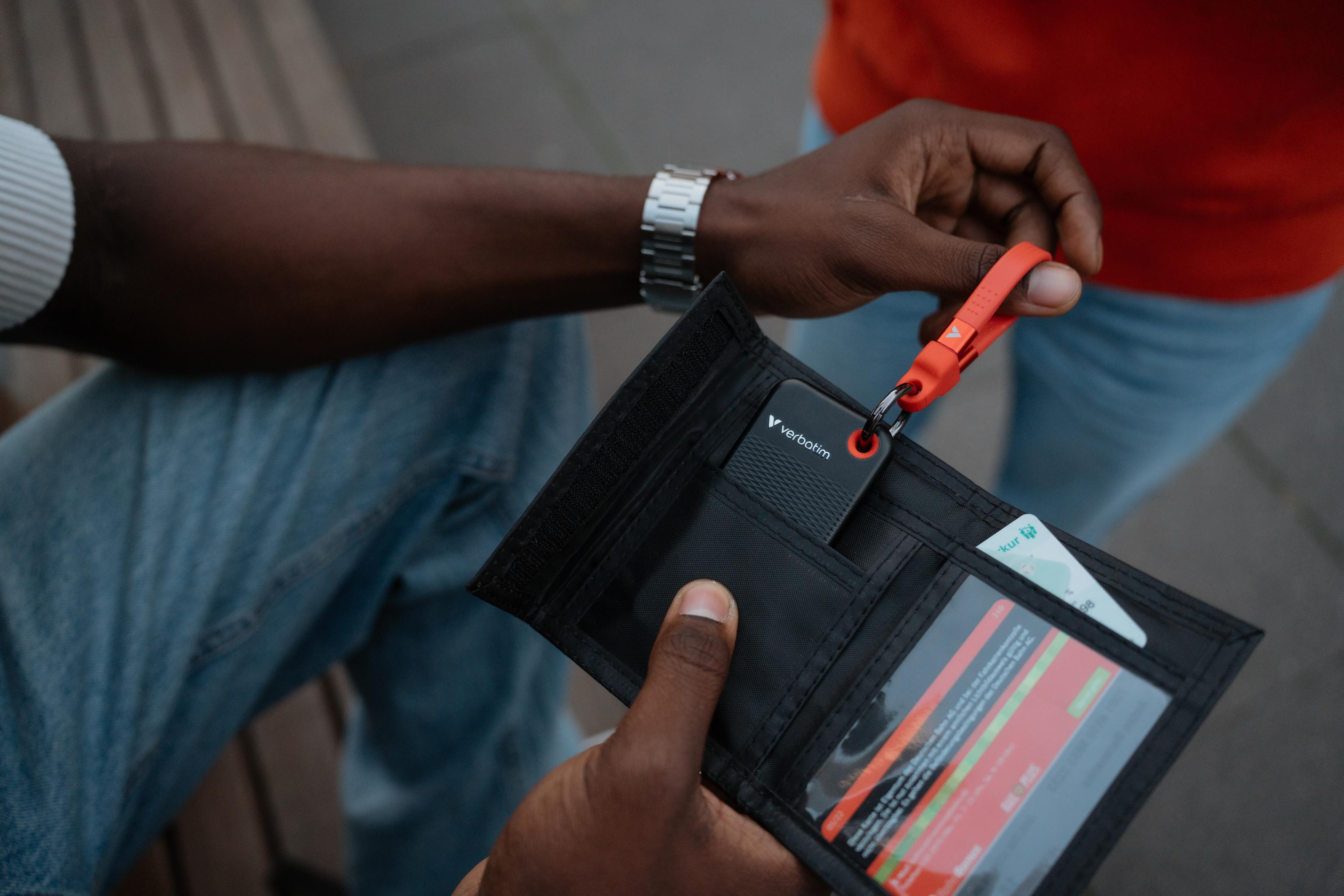 Osoba trzymająca czarny portfel Verbatim z czerwonym paskiem, w pobliżu drugiej osoby.