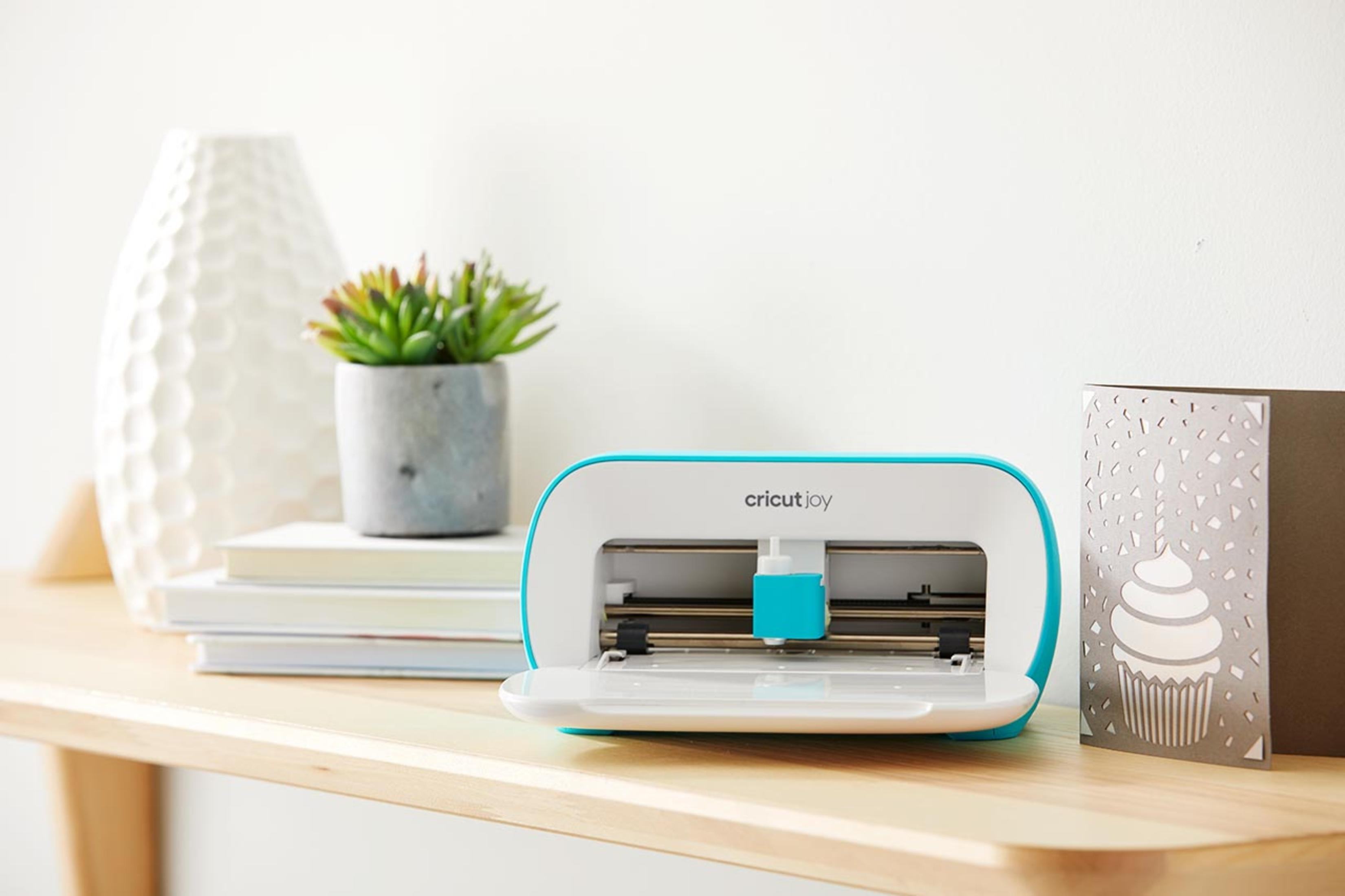 Türkis-weiße Cricut Joy Maschine mit Karte und kleiner Sukkulente auf einem hellen Holztisch.