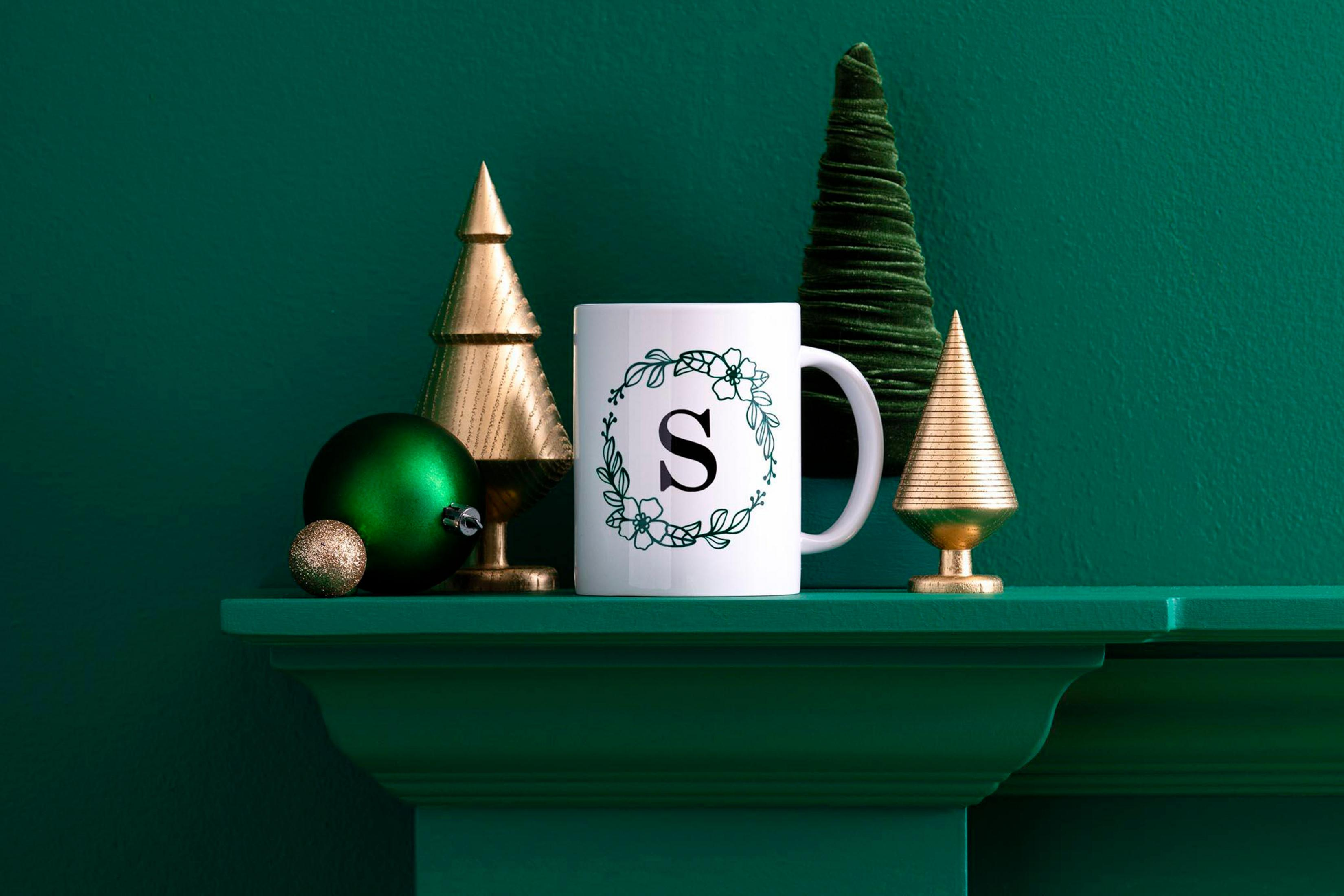 Eine weiße Tasse mit Blumengirlande und Initialen auf einem Kaminsims mit Weihnachtsdeko. Grüner Hintergrund.