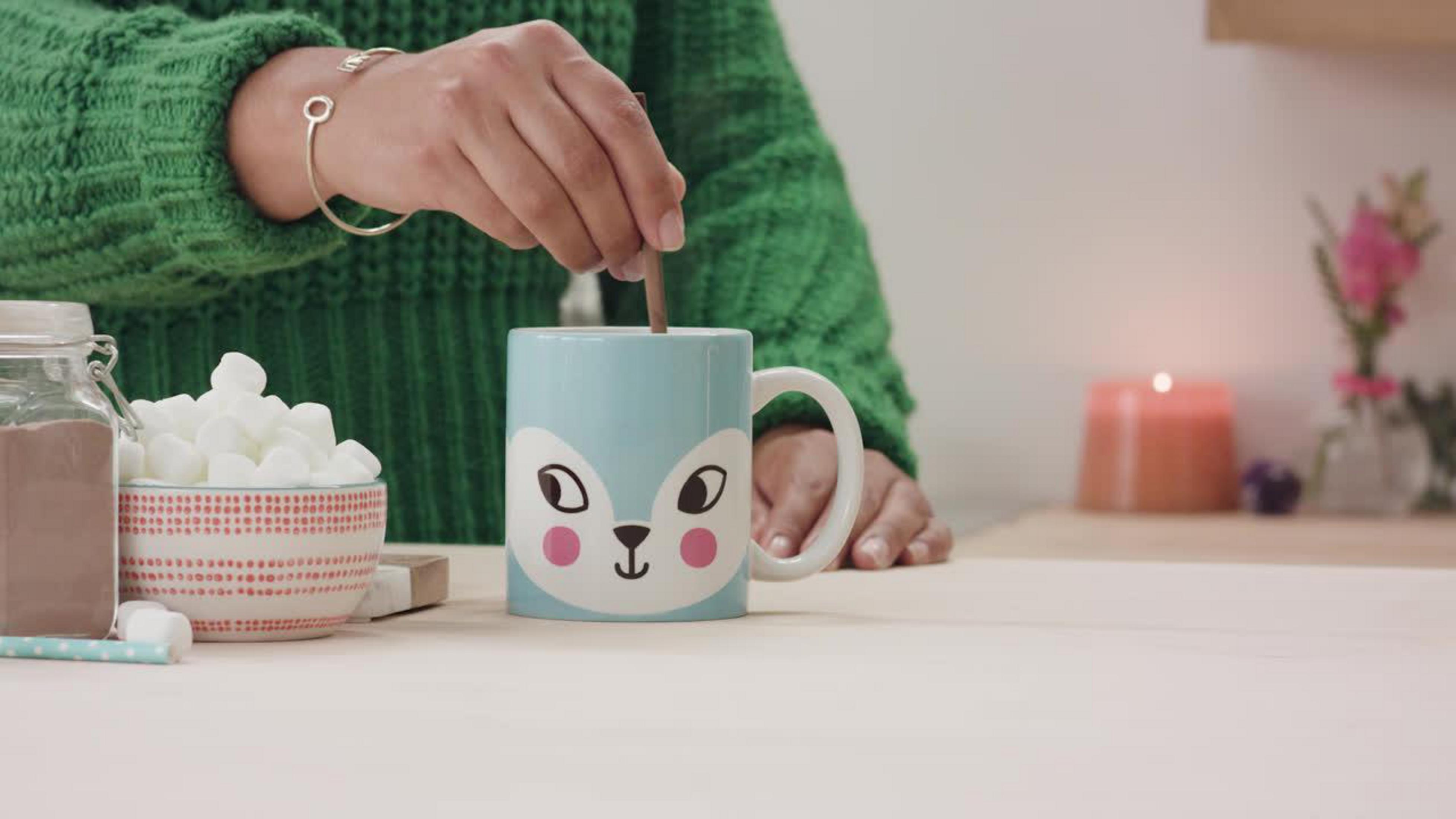 Person rührt in einer Tasse mit Fuchsmotiv, Marshmallows und Kakao daneben.