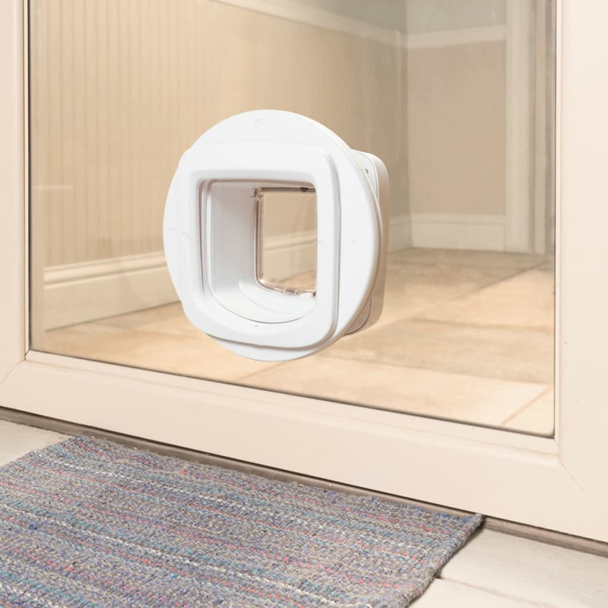 Eine weiße Haustiertür, die in einem Glasfenster installiert ist. Unter der Tür liegt ein blau-grauer Teppich.