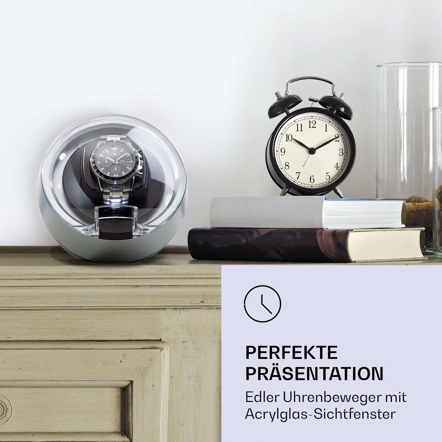 Eine Uhr in einer durchsichtigen Vitrine auf einer Holzoberfläche, neben einem schwarzen Wecker und Büchern.