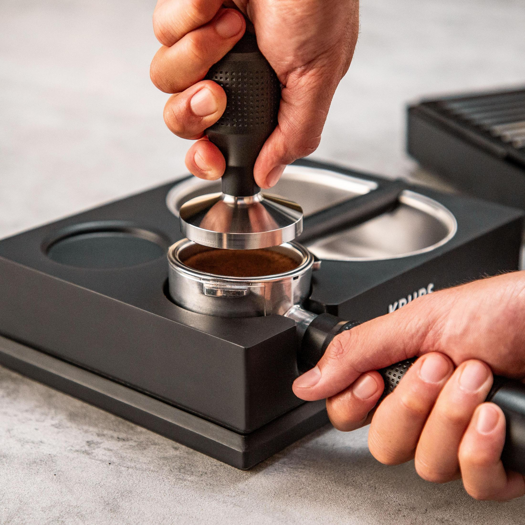 Ręce używające ubijaka do wciskania zmielonej kawy w kolbę na czarnym stojaku.