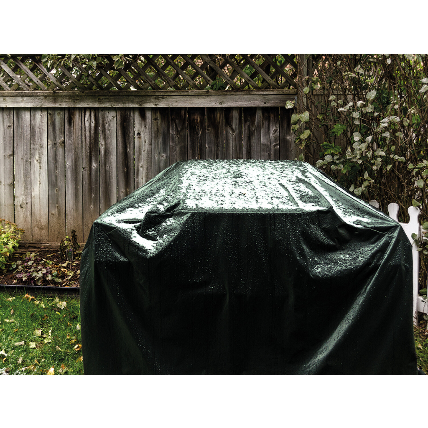 Ciemnozielona osłona grilla jest na grillu. Tło przedstawia drewniany płot i zieleń.