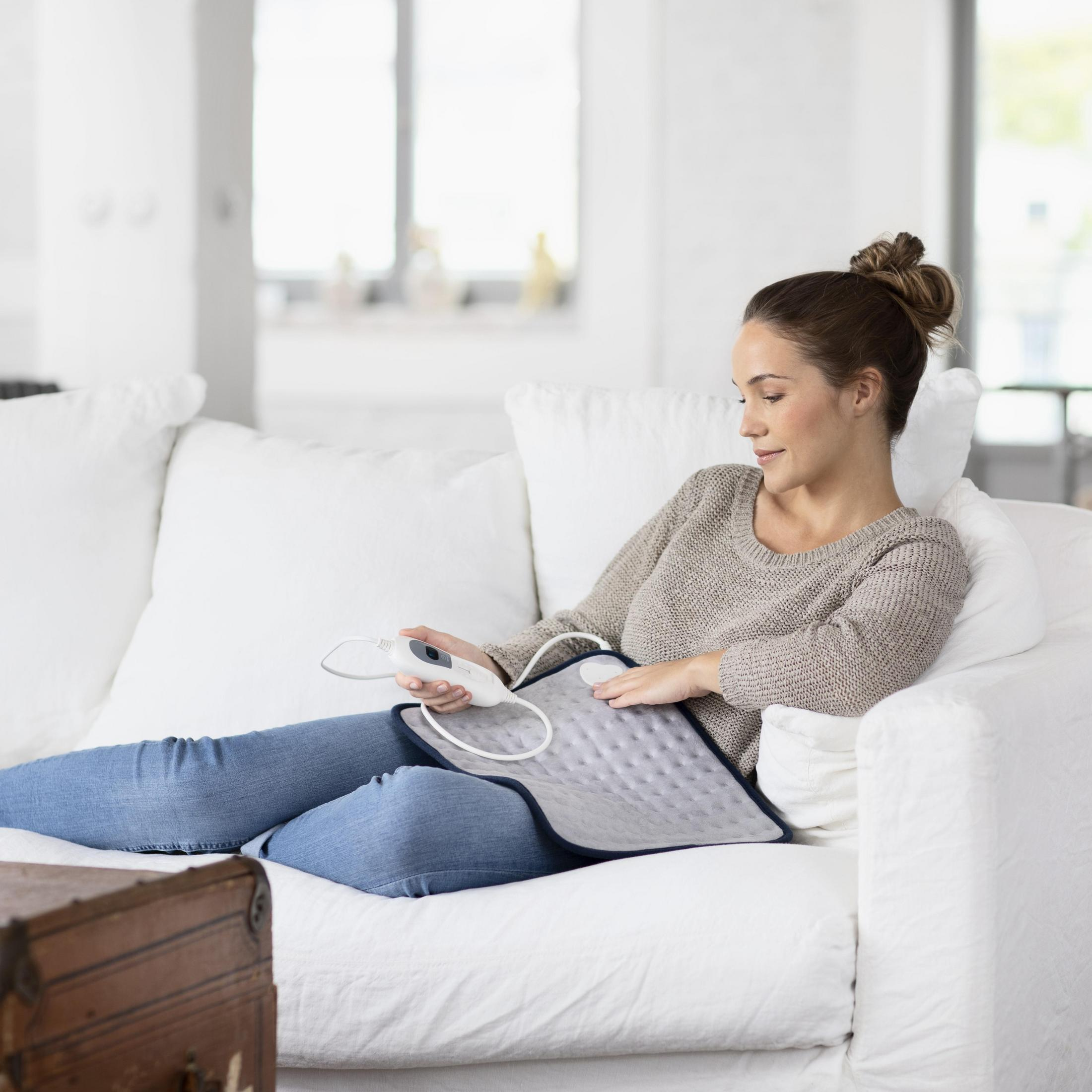 Kobieta na białej sofie z podkładką grzewczą, w dżinsach i swetrze. Drewniana skrzynia w tle.