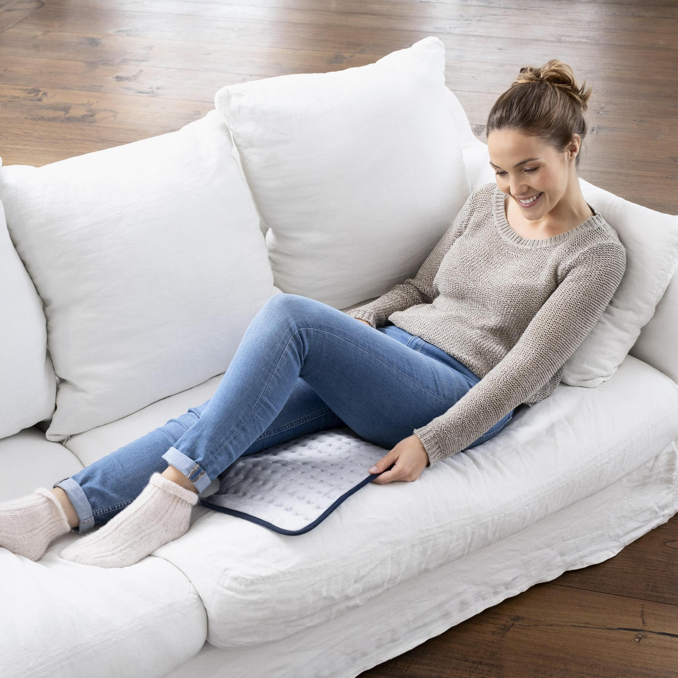 Kobieta na białej sofie, używająca podkładki grzewczej. Ma na sobie dżinsy i sweter. Przytulna scena.