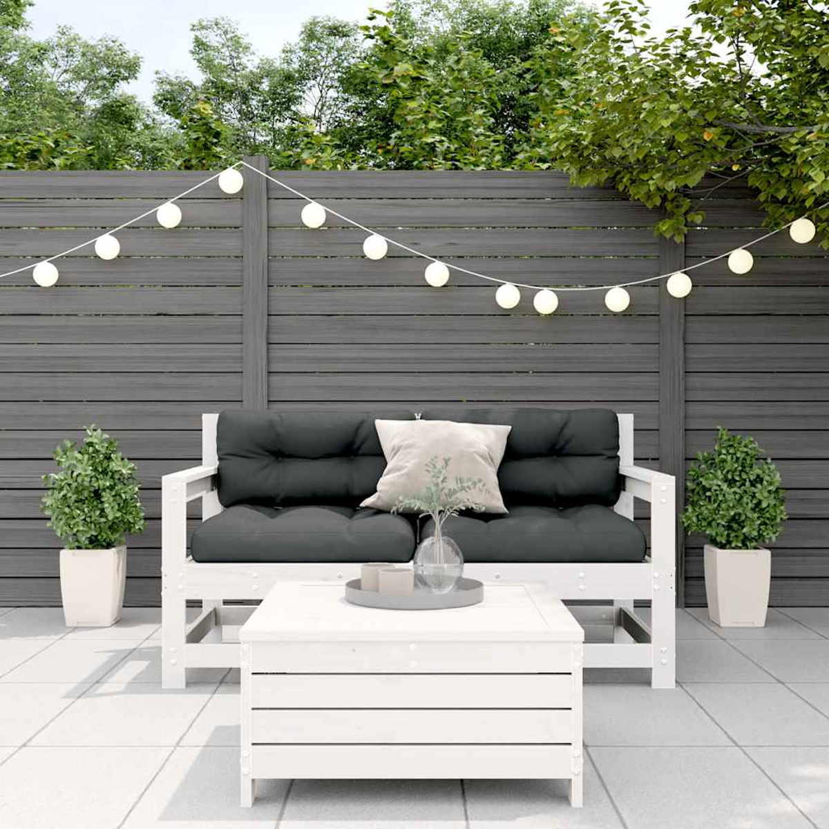 Weißes Outdoor-Sofa mit schwarzen Kissen, einem weißen Tisch und Pflanzen auf einer Terrasse mit Lichtern.