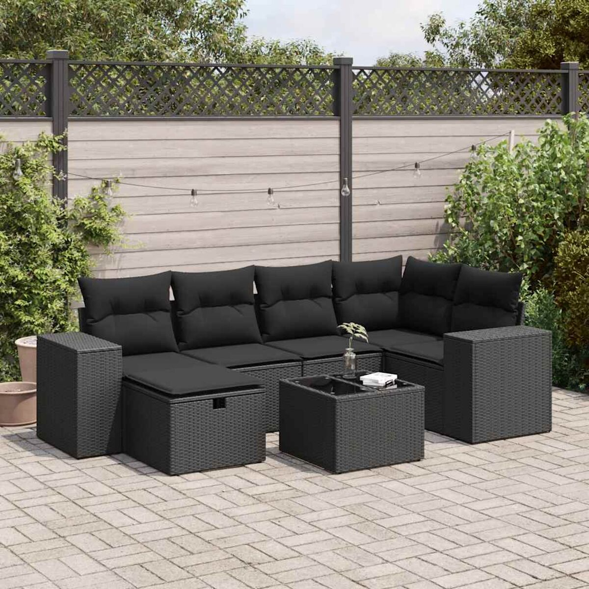 Schwarzes Korbmöbel-Ecksofa-Set auf einer gepflasterten Terrasse. Schwarze Kissen und grauer Zaun im Hintergrund.