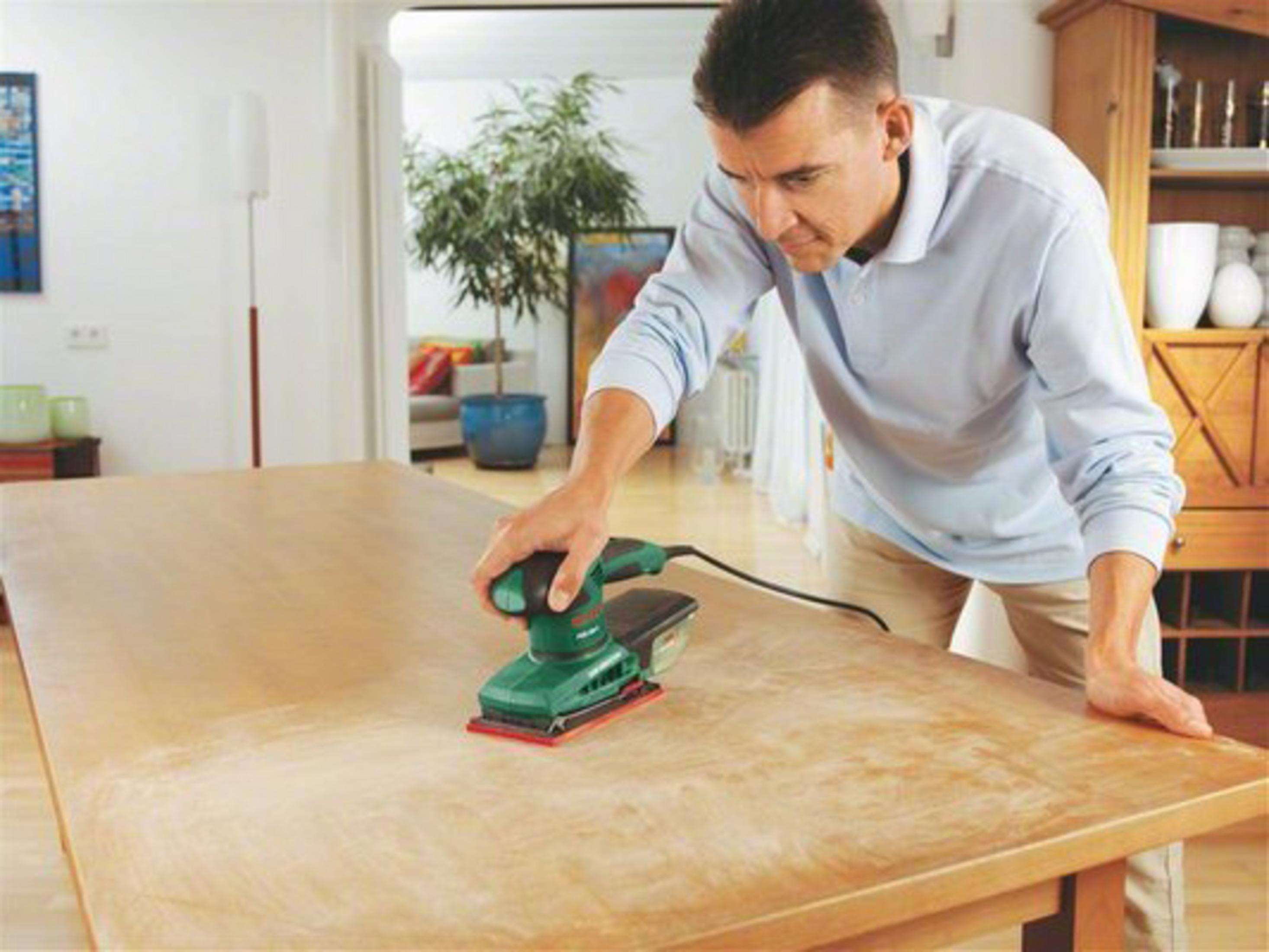 Homme ponçant une table en bois avec une ponceuse verte et noire.