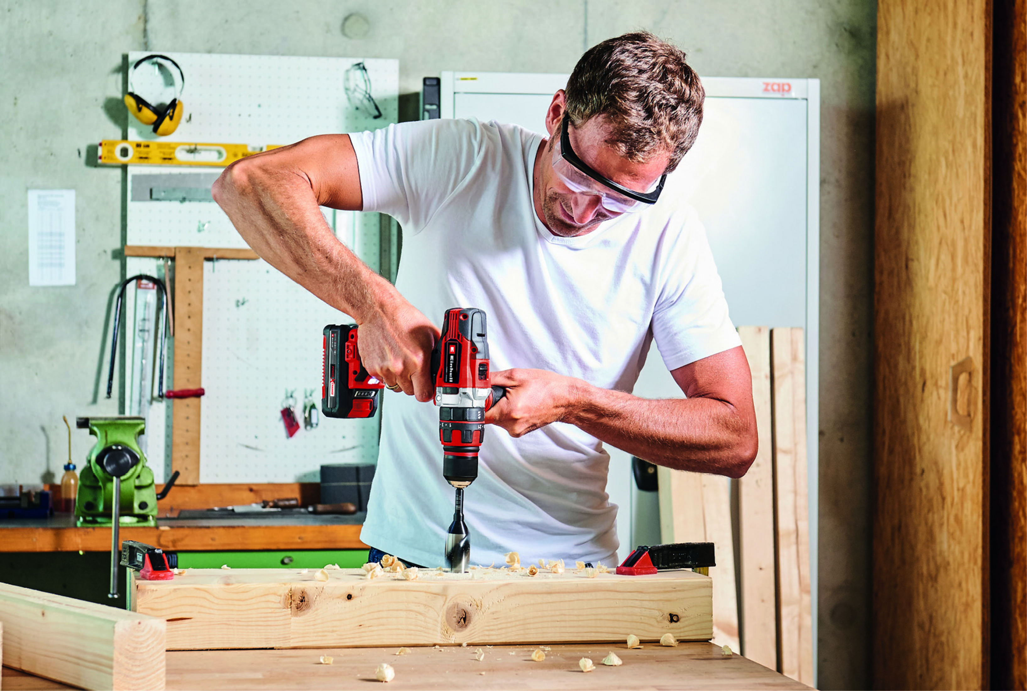 Mężczyzna w okularach ochronnych używający czerwono-czarnej wiertarki na drewnianej desce. Warsztat.