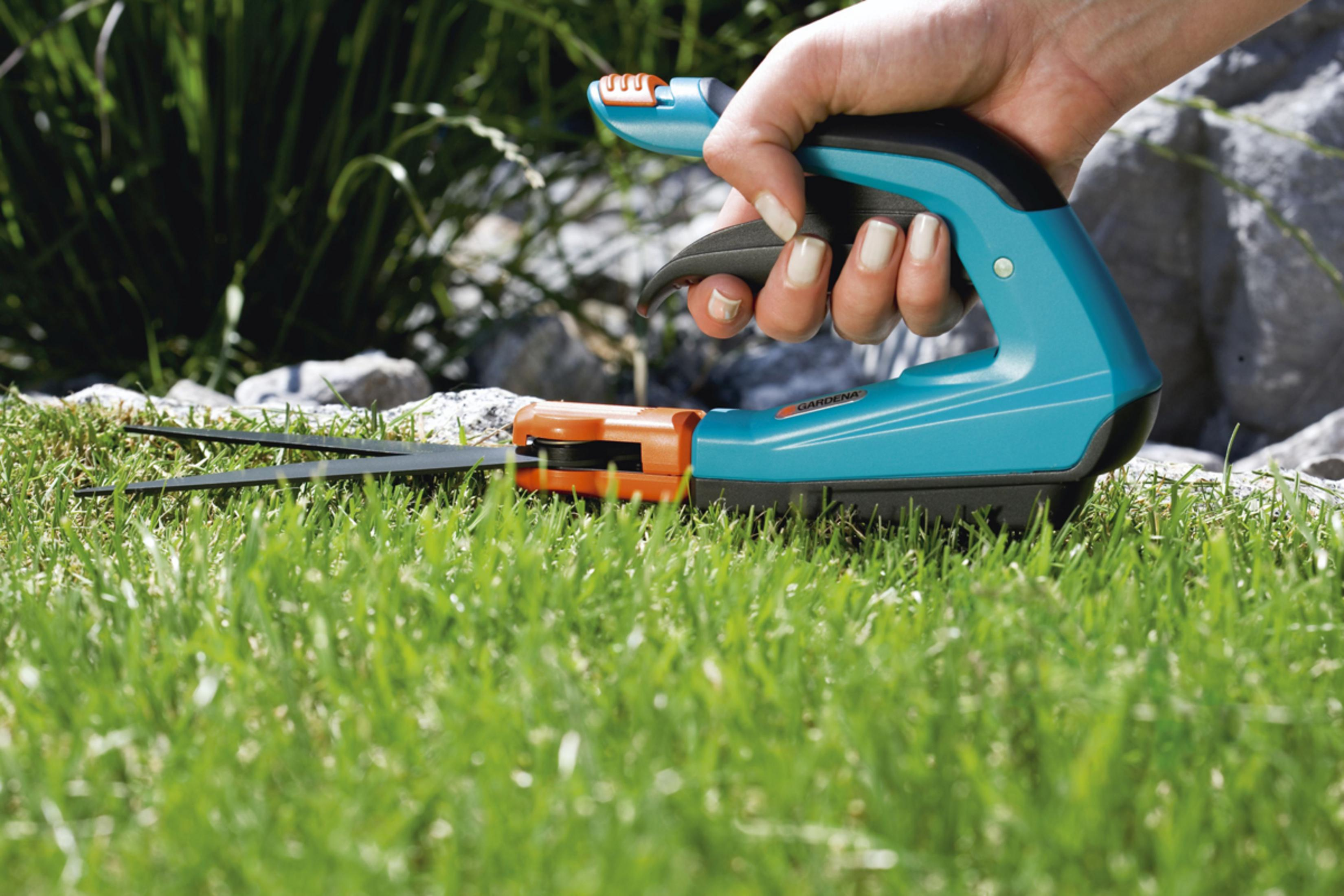 Dłoń trzymająca zielono-pomarańczowe nożyce do trawy. Zielona trawa i skały w tle.