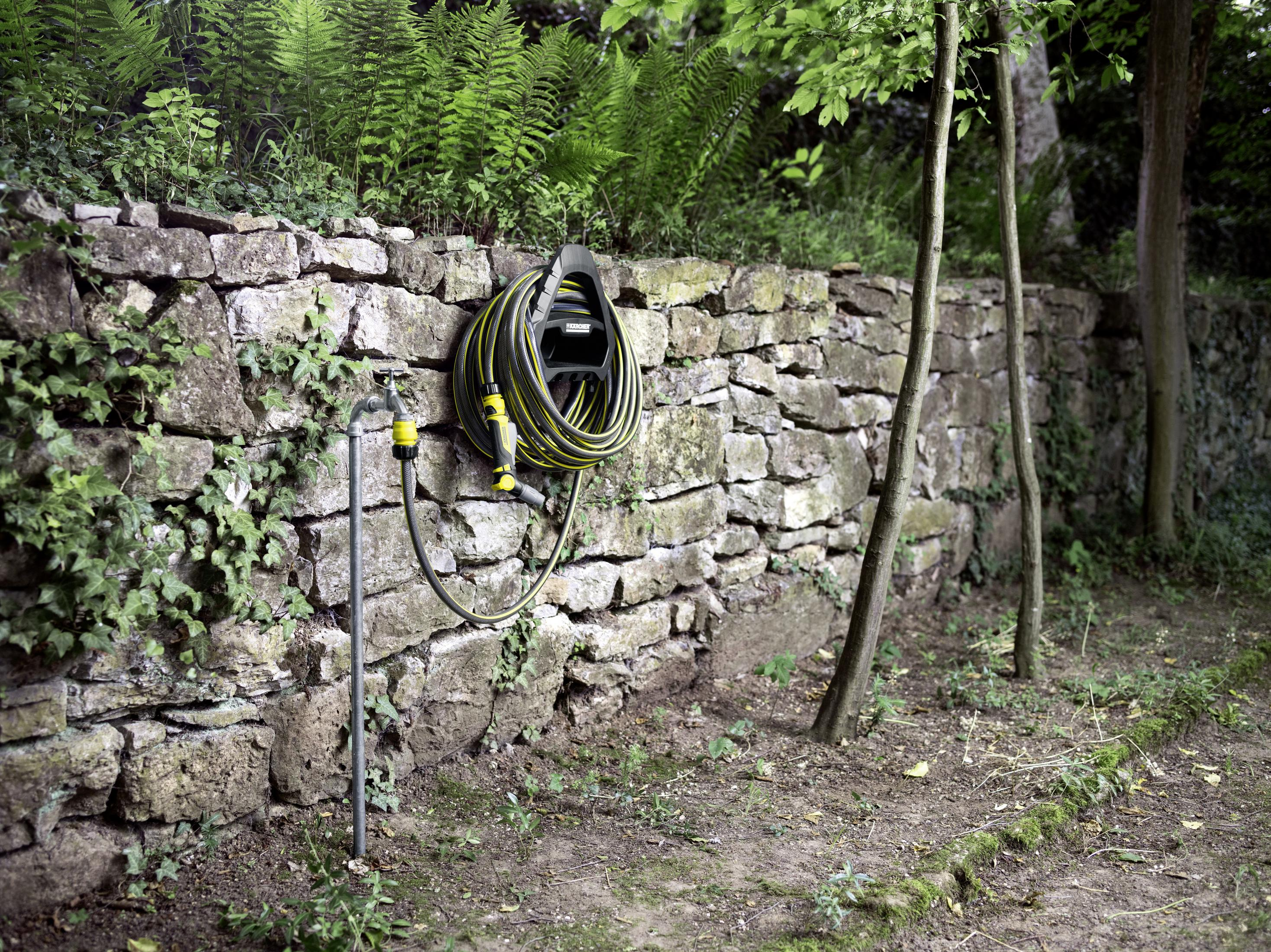Schlauchtrommel an einer Steinmauer. Ein schwarzer Schlauch ist an Wasserhahn und Trommel angeschlossen. Grüne Pflanzen im Hintergrund.