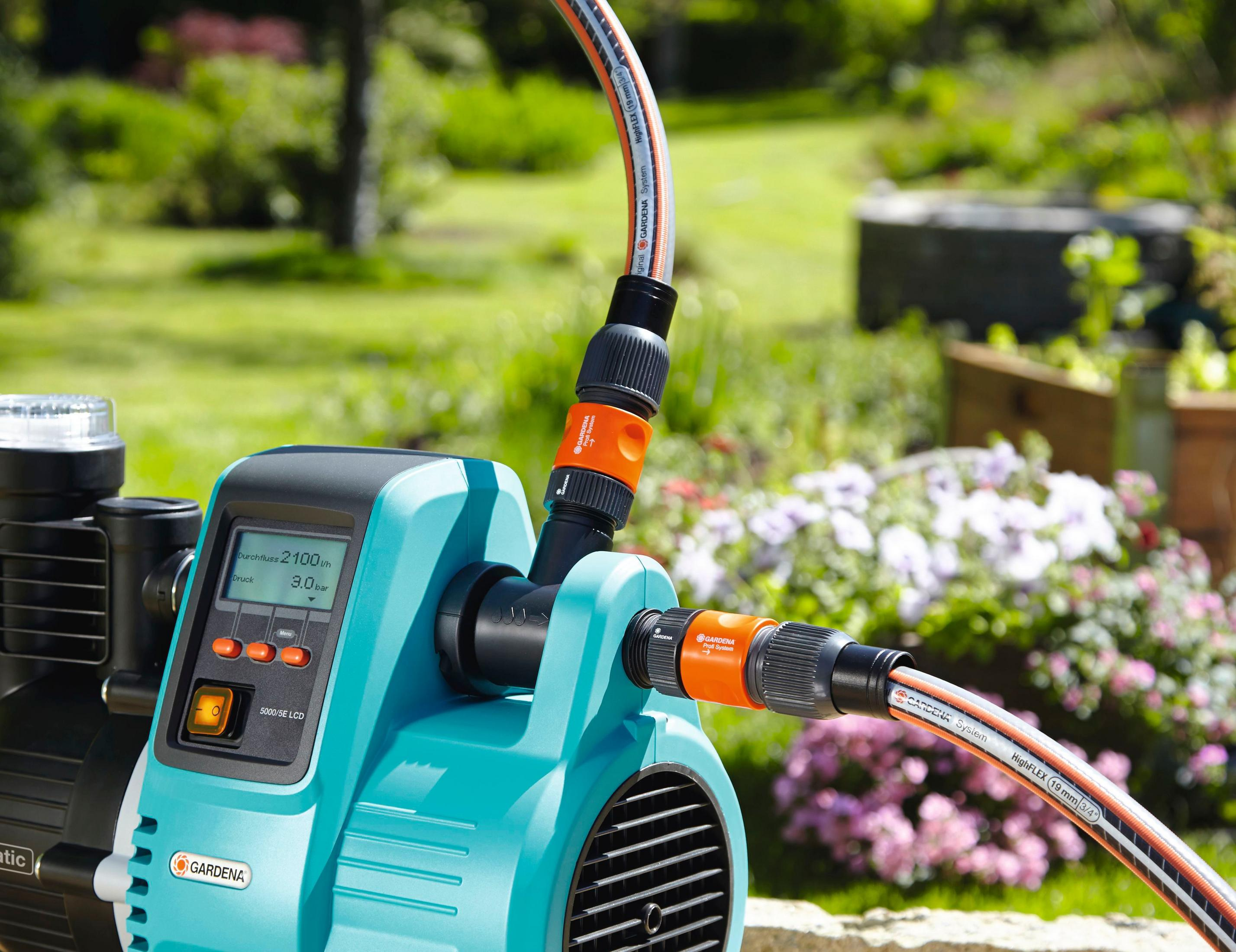 Nahaufnahme einer türkisblauen Wasserpumpe mit angeschlossenen Schläuchen und Garten im unscharfen Hintergrund.