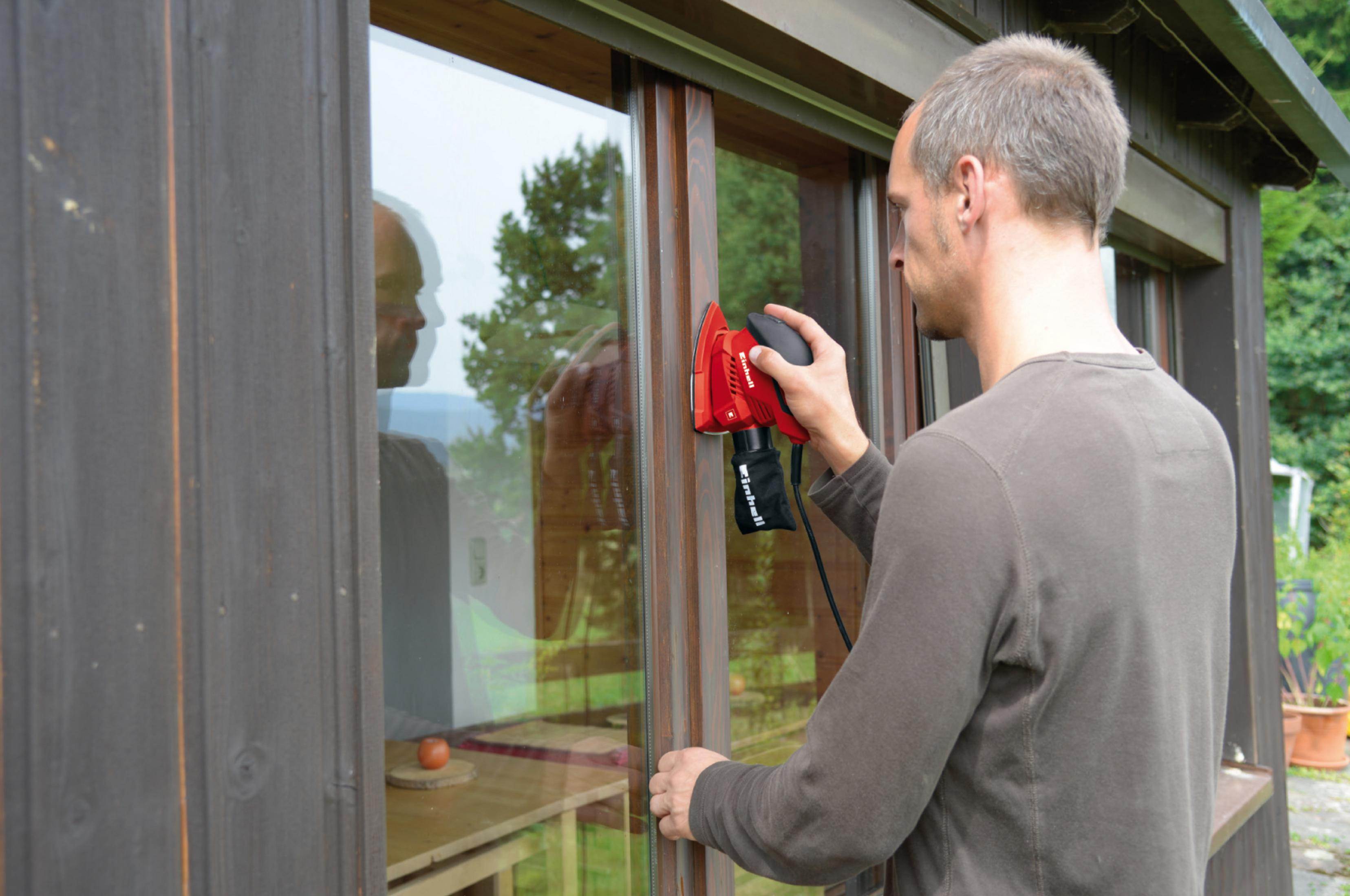 Mężczyzna szlifuje ramę okienną czerwoną szlifierką mimośrodową. Odbicie w oknie.