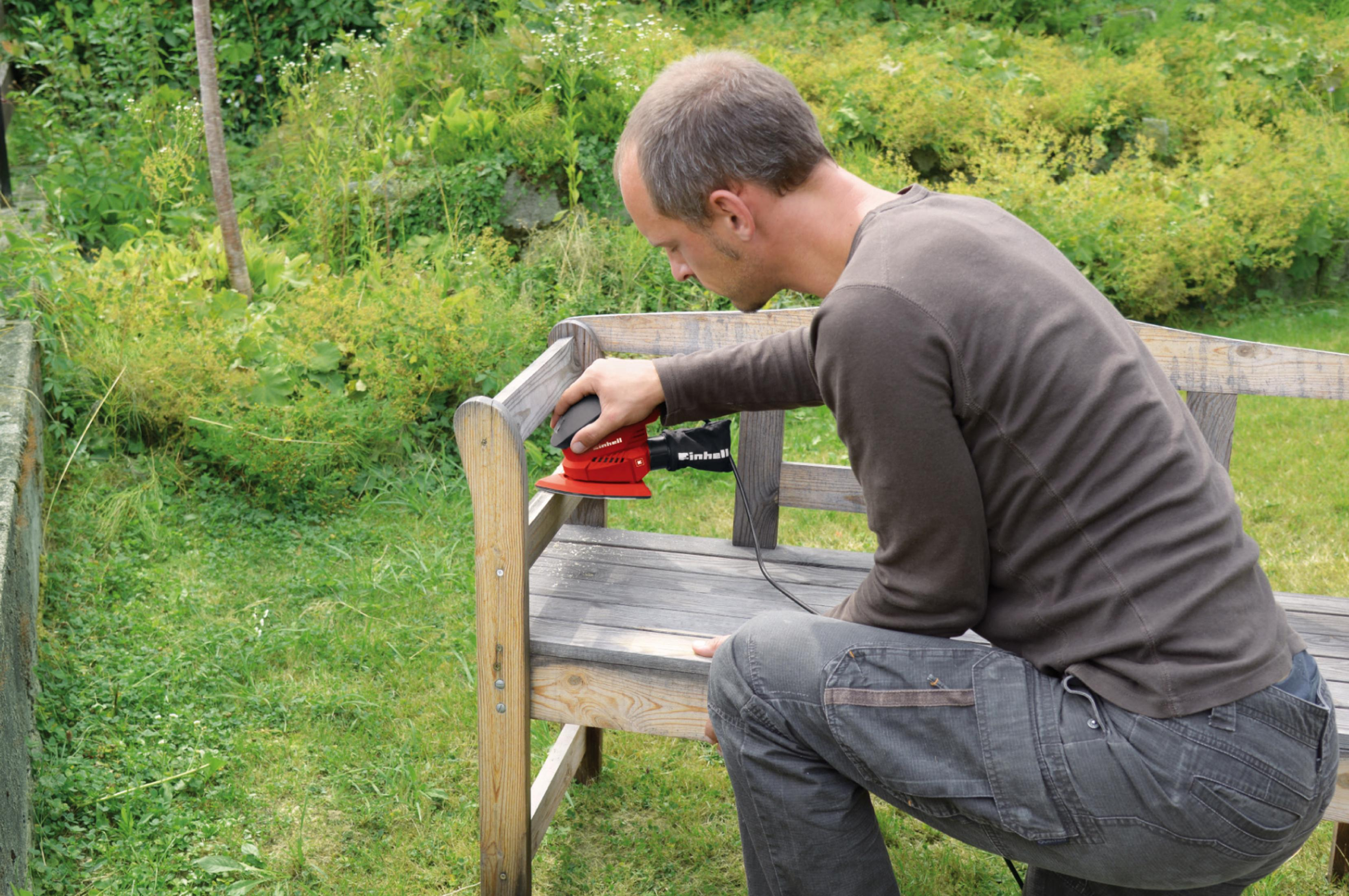 Mężczyzna szlifuje drewnianą ławkę szlifierką elektryczną. Zielona trawa i krzewy w tle.