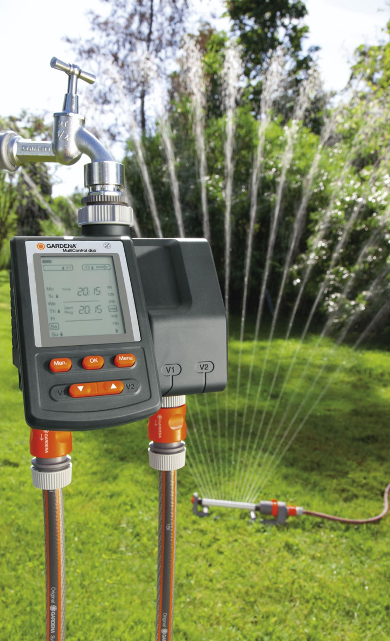 Un dispositivo Gardena water control duo su un prato verde. L'acqua spruzza da un irrigatore sullo sfondo.