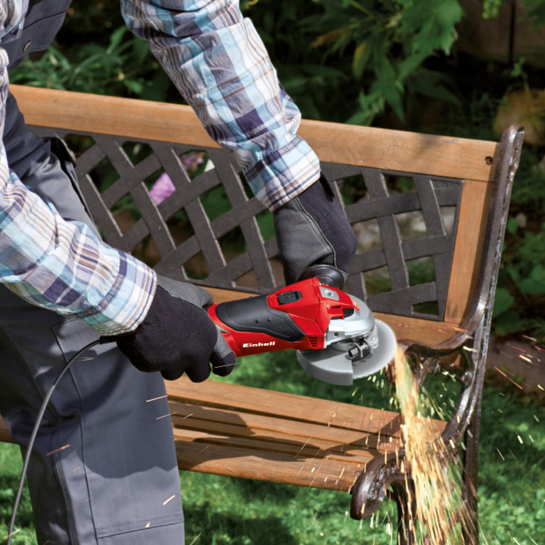 Una persona usando una amoladora angular roja para cortar un banco de madera marrón. Saltan chispas.