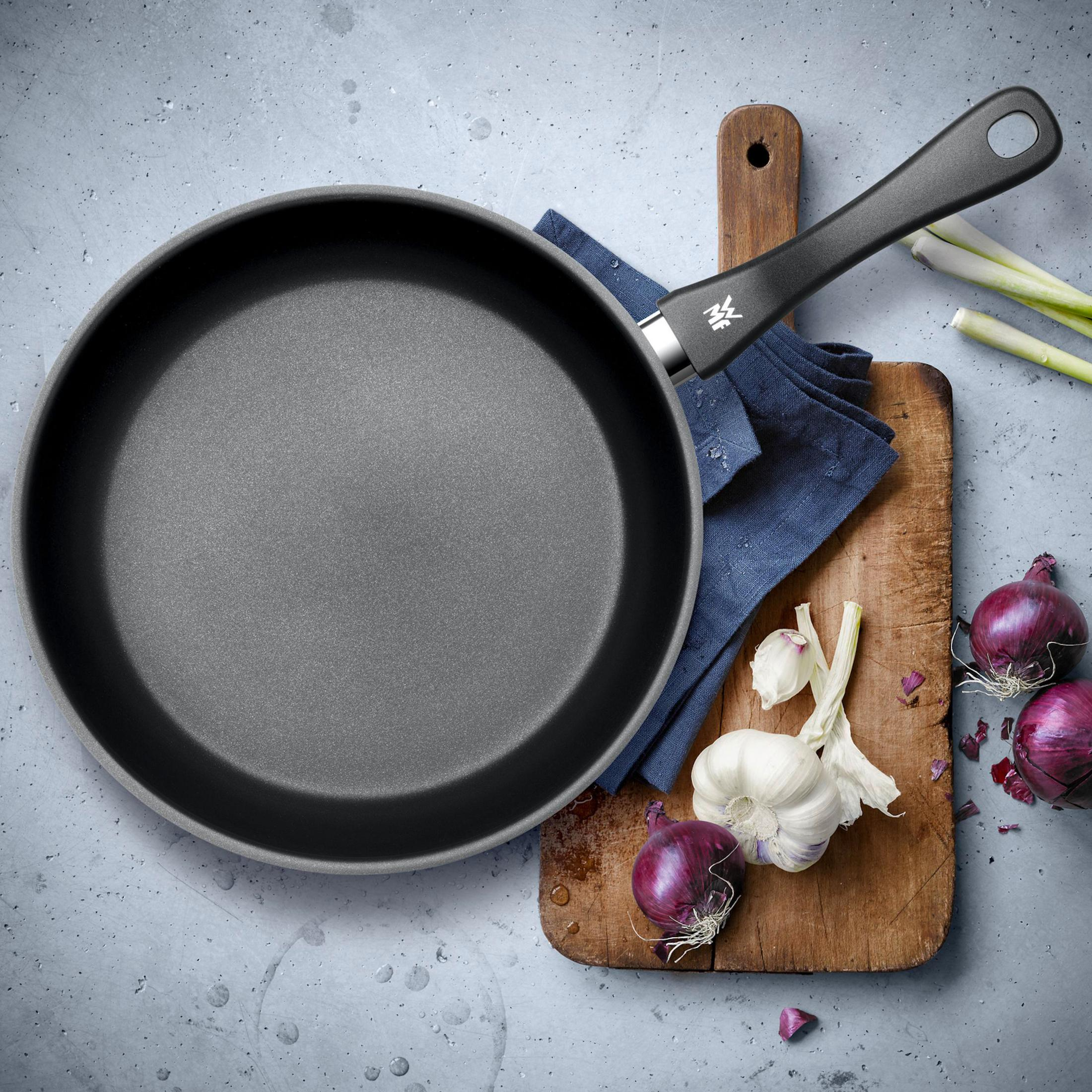 Widok z góry na patelnię ze składnikami. Zawiera cebulę, czosnek, niebieską szmatkę i drewnianą deskę.