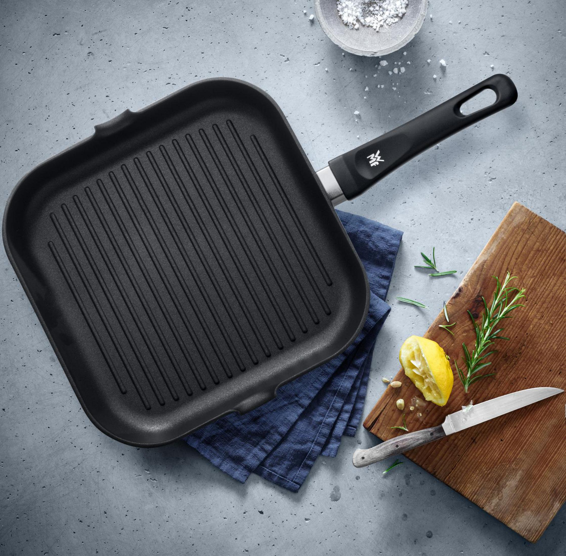 Schwarze Grillpfanne mit Griff, Zitrone, Rosmarin und Messer auf Steinoberfläche.