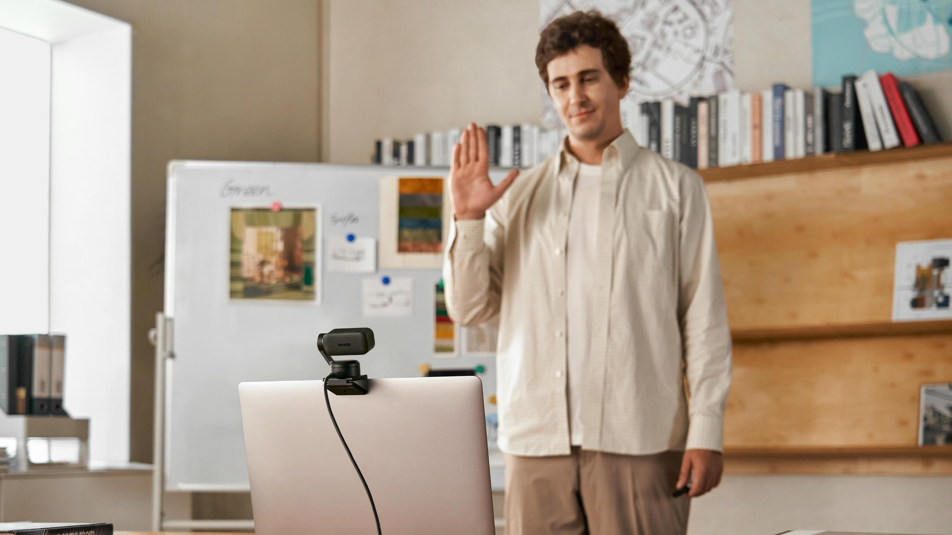 Mężczyzna jest na rozmowie wideo. Stoi przed tablicą, macha, z laptopem i kamerą internetową.