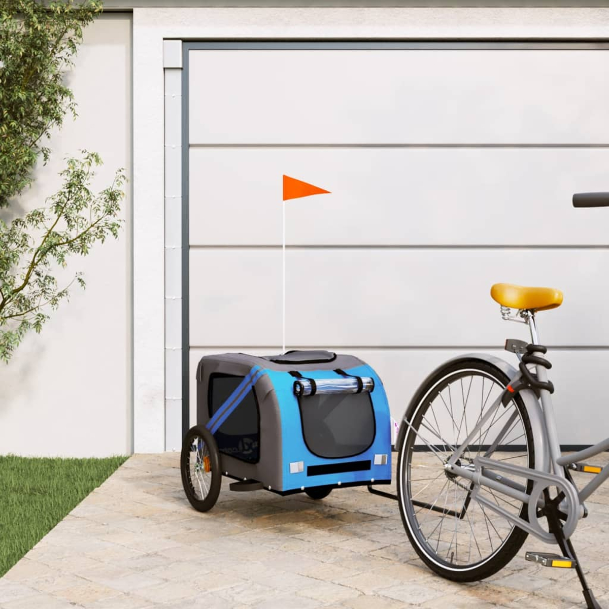 Ein blau-grauer Haustieranhänger ist an einem Fahrrad befestigt. Eine orangefarbene Fahne ist befestigt. Weißes Garagentor.