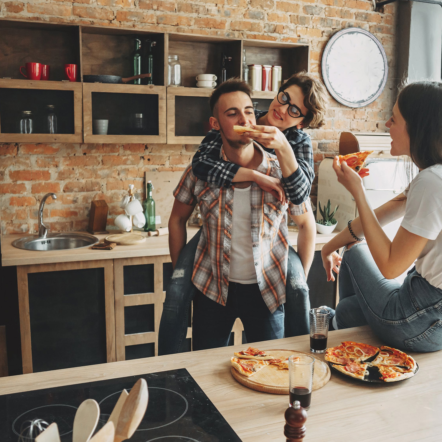 Drei Personen in einer Küche. Ein Mann wird von einer Frau mit Pizza gefüttert. Eine andere Frau isst Pizza.