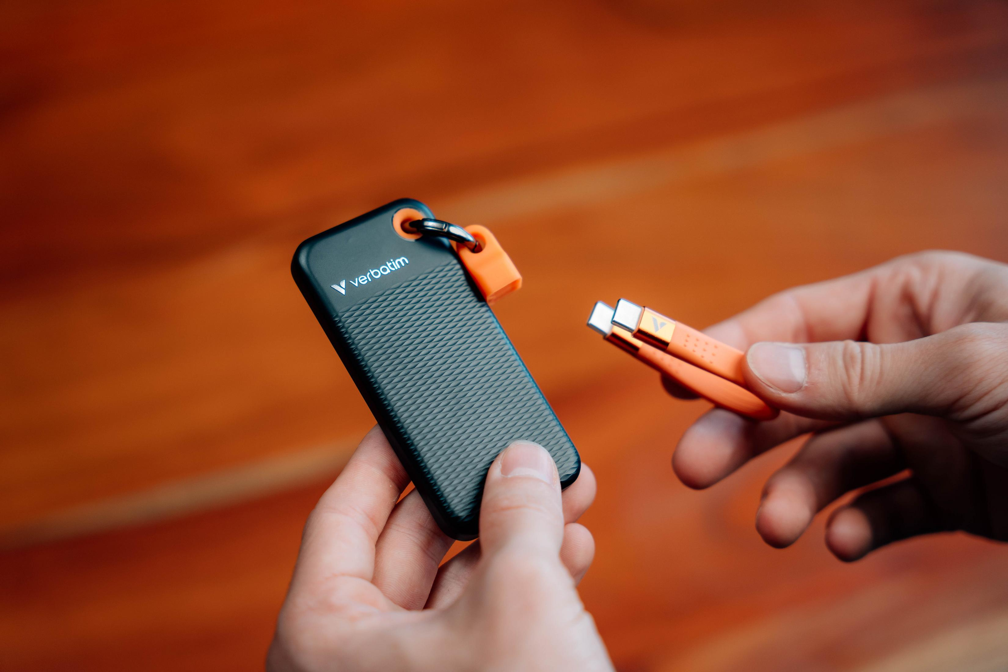 Dłonie trzymają czarny dysk SSD Verbatim i pomarańczowe mocowanie kabla. Drewniane tło.