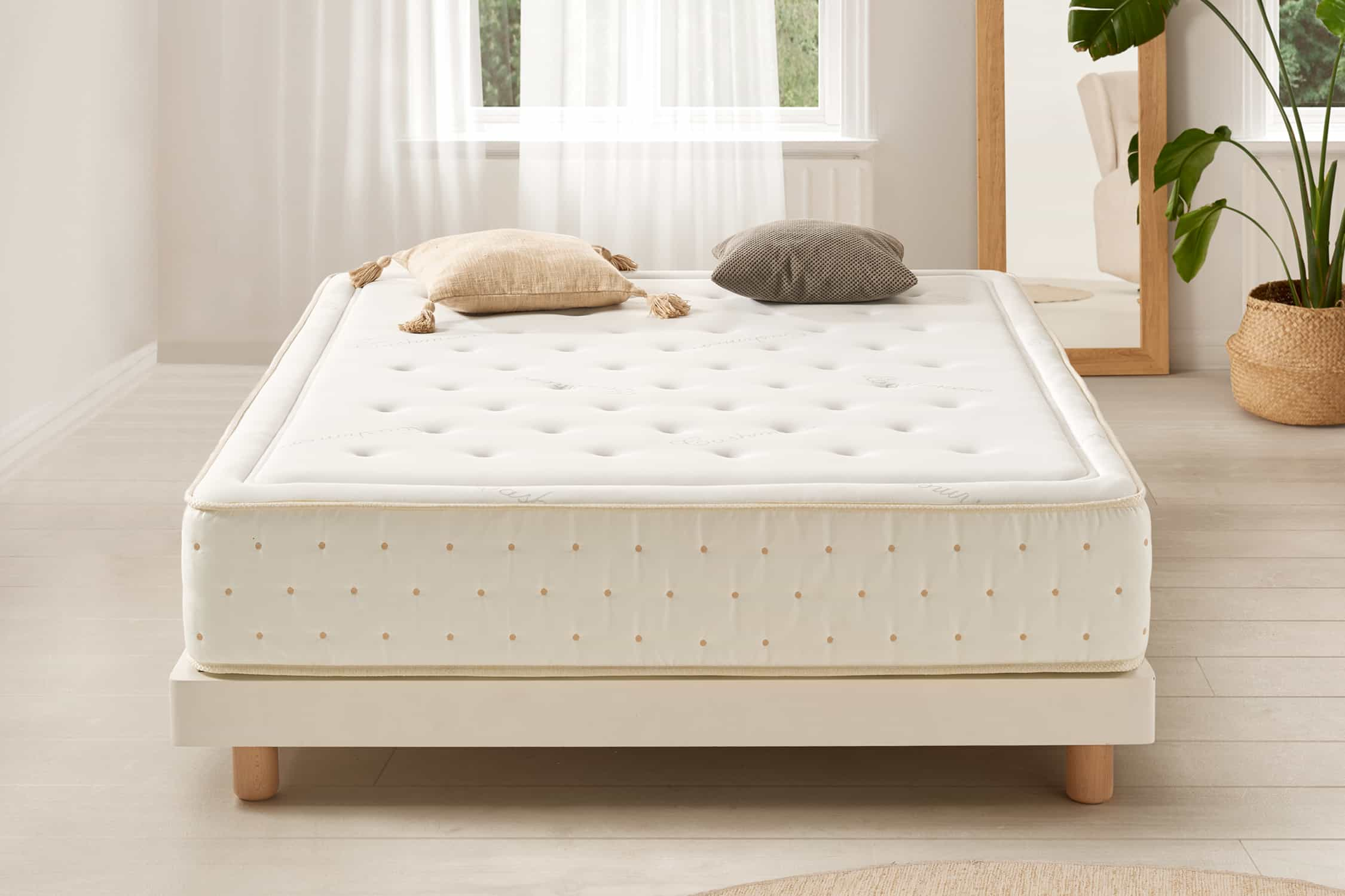 Habitación con cama con colchón crema, almohadas beige y grises, y muebles de madera. Ventana al fondo.