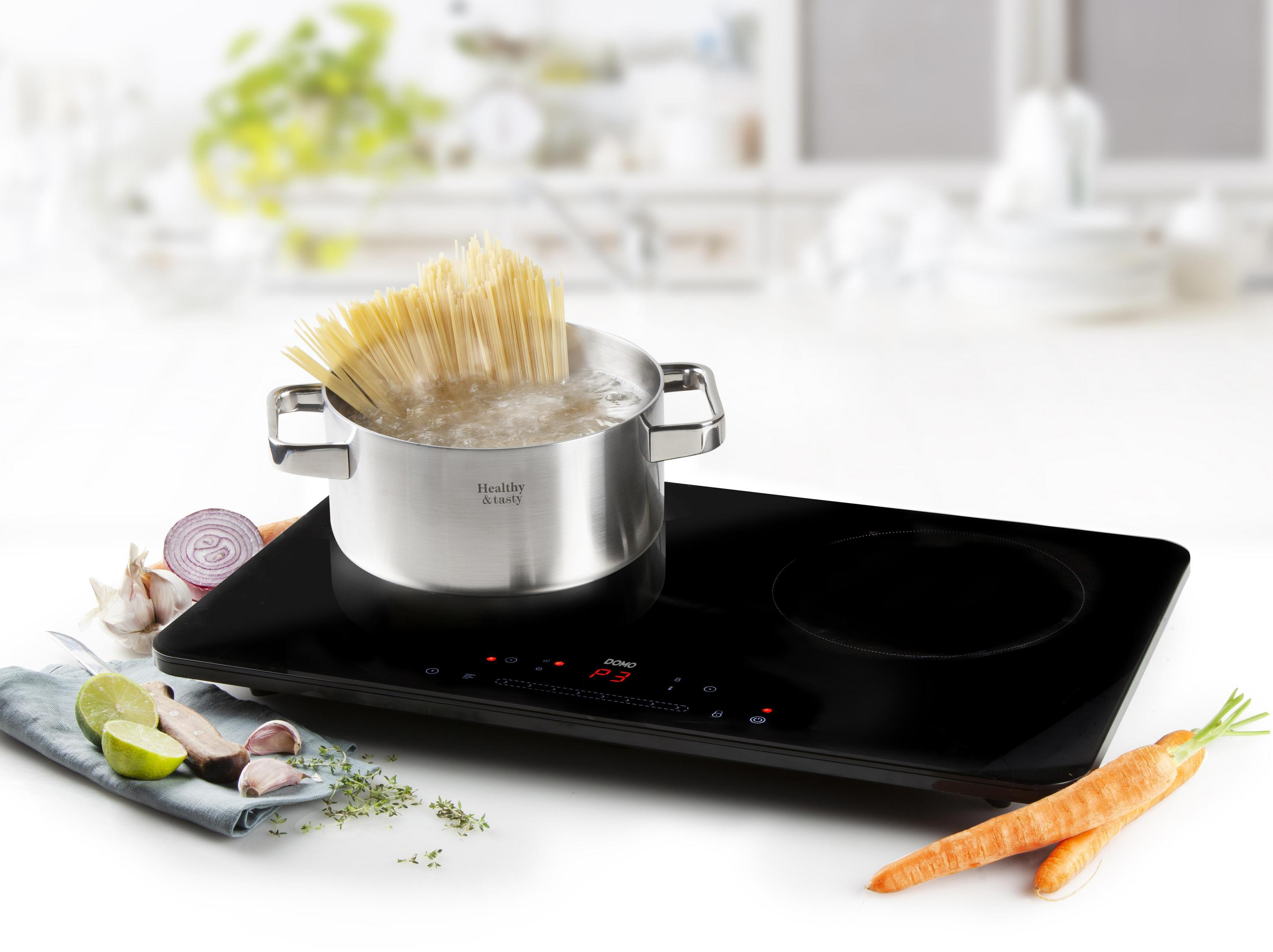 Plaque à induction avec casserole de pâtes bouillantes. Des ingrédients comme carottes, ail et herbes sont placés autour.