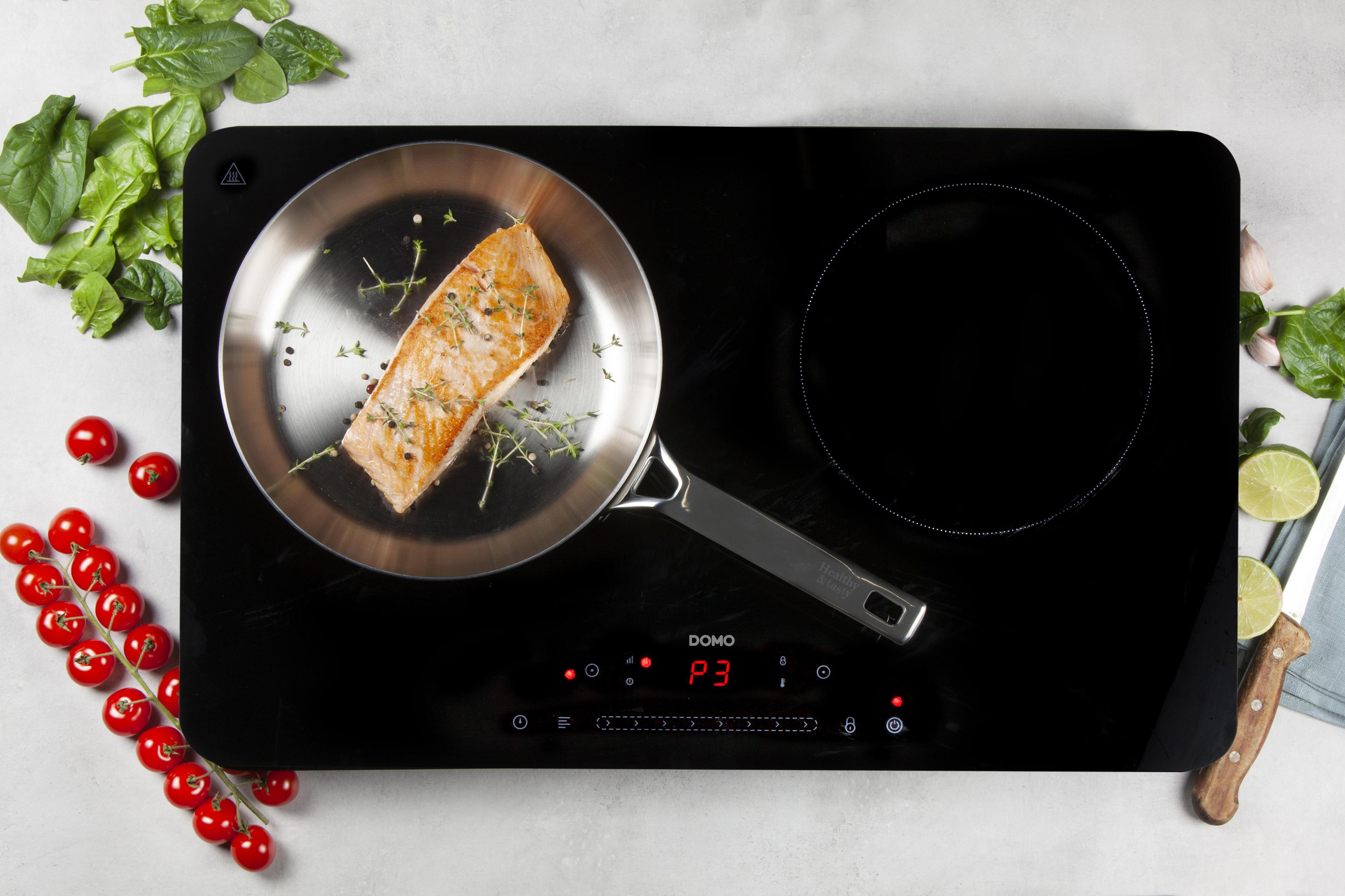 Vue aérienne d'une table de cuisson à induction noire avec une poêle de saumon et légumes.