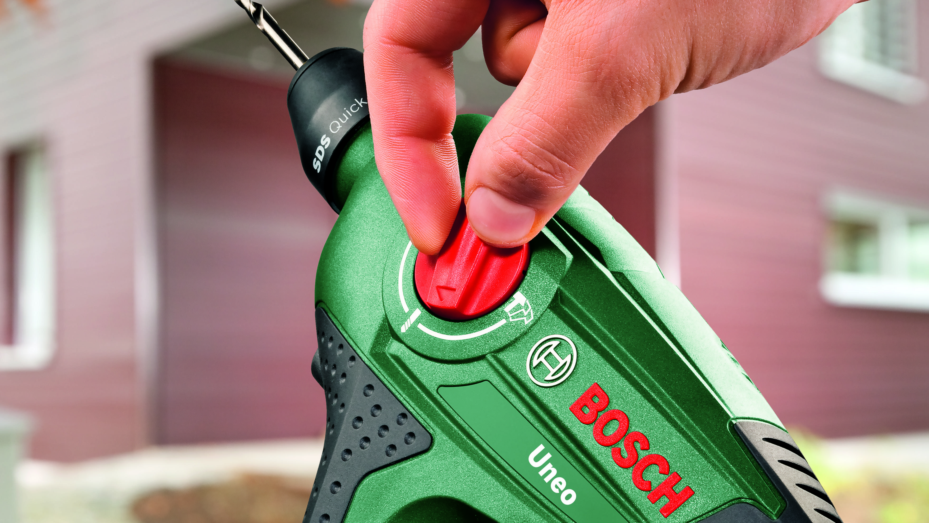 Ręka naciska czerwony przycisk na zielonej wiertarce Bosch z wiertłem.