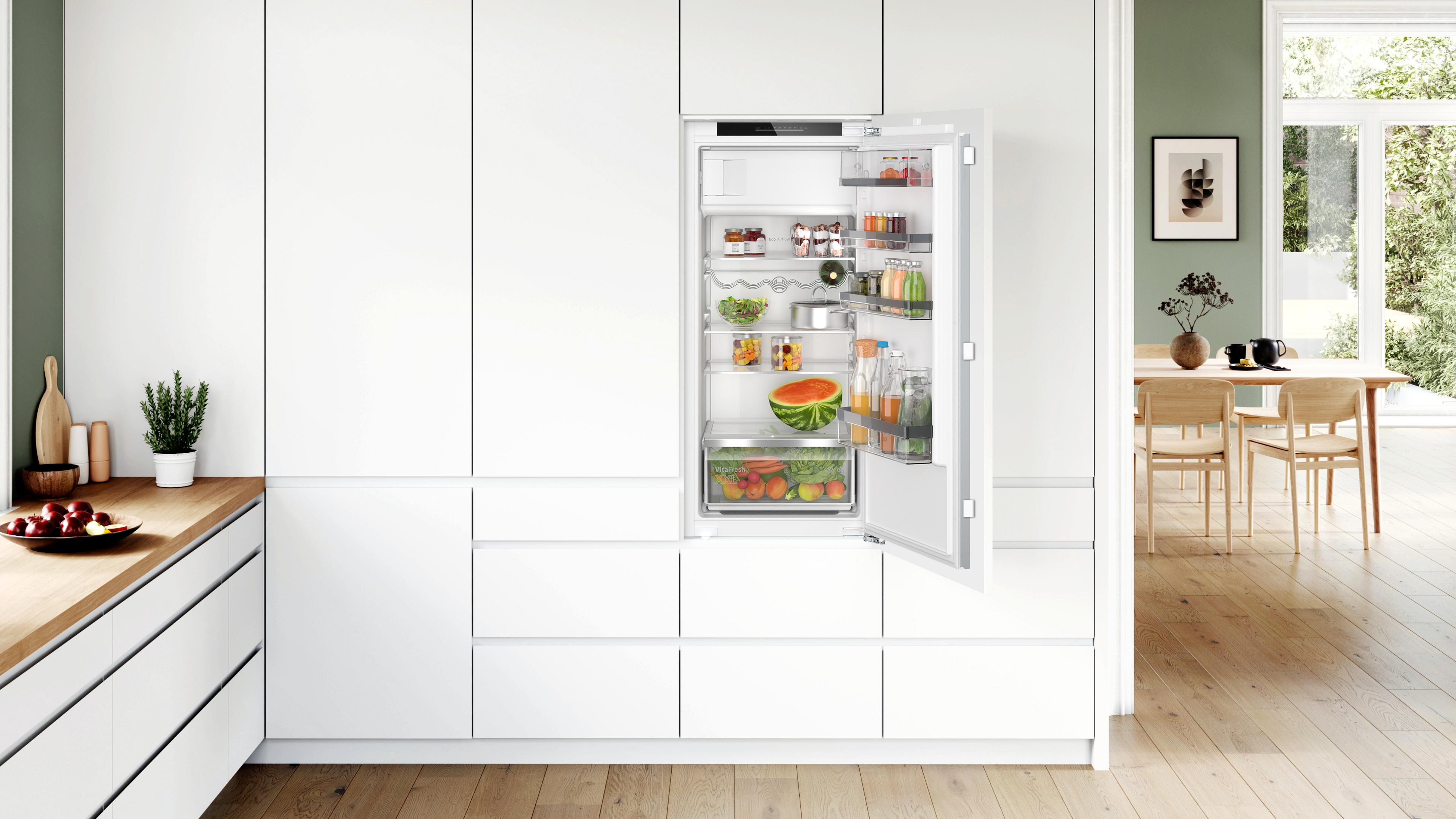 Weißer Kühlschrank mit geöffneter Tür, die Regale mit Lebensmitteln zeigt. Die Küche ist weiß mit Holz.