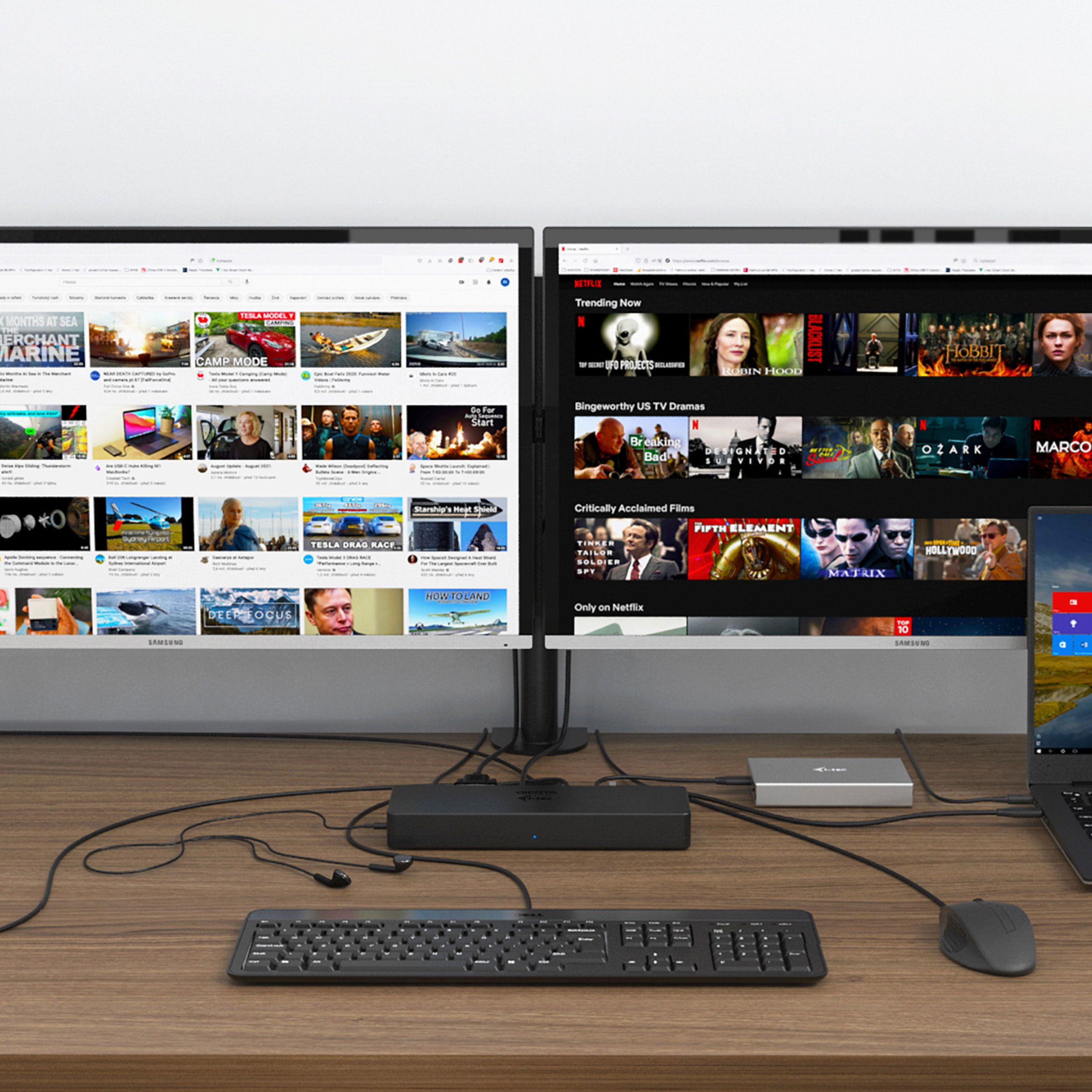 Due monitor di computer mostrano contenuti. Tastiera, mouse e hub visibili su una scrivania in legno.