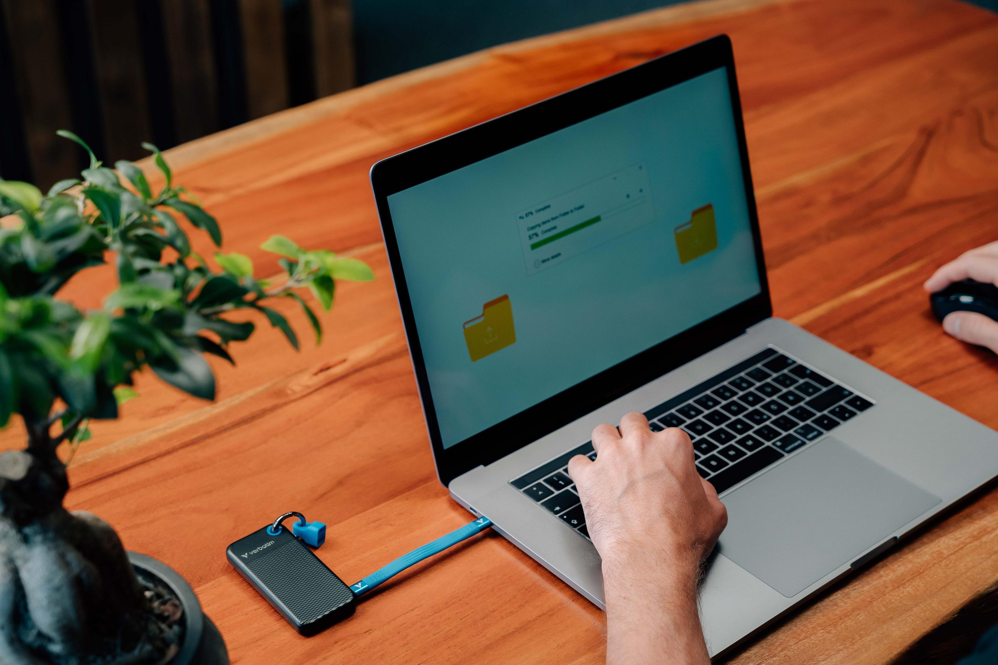 Laptop z podłączonym urządzeniem Verbatim, pokazuje transfer plików. Ręka używa myszy.