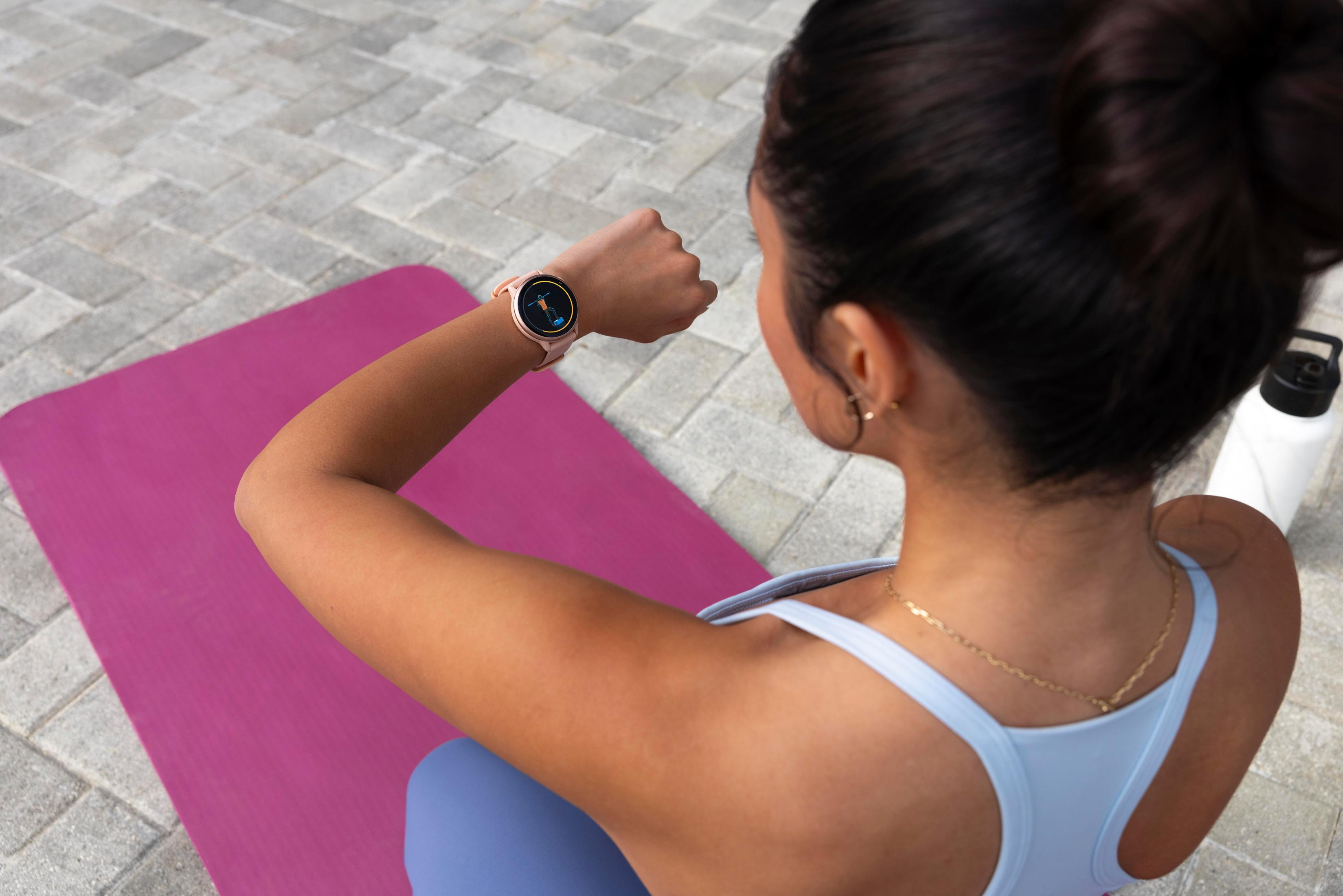 Mujer con smartwatch, mirándolo mientras hace ejercicio en una esterilla rosa, al aire libre.
