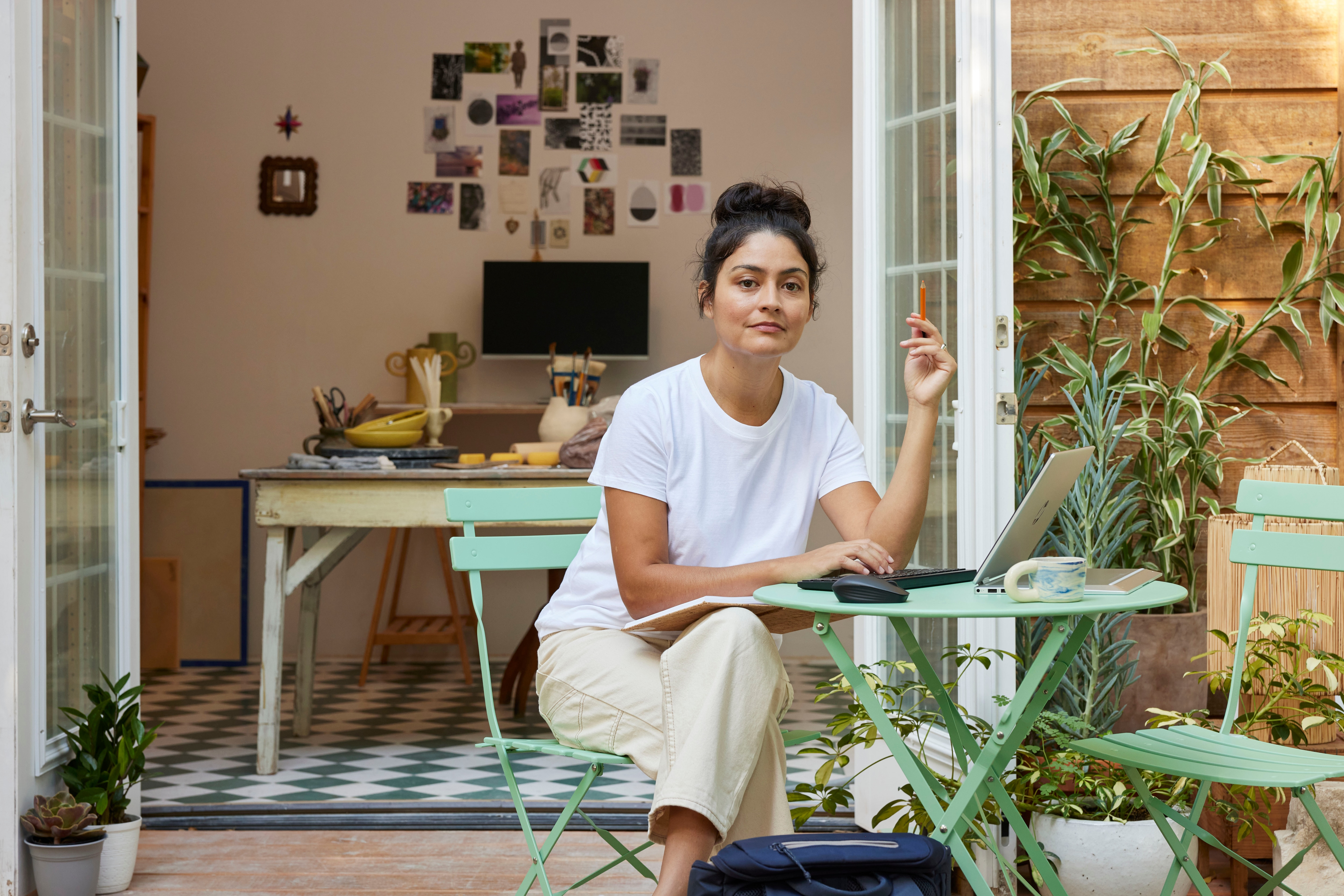Kobieta w białej bluzce siedzi przy stole pracując na laptopie, w ogrodzie. Widoczne zielone krzesła i rośliny.
