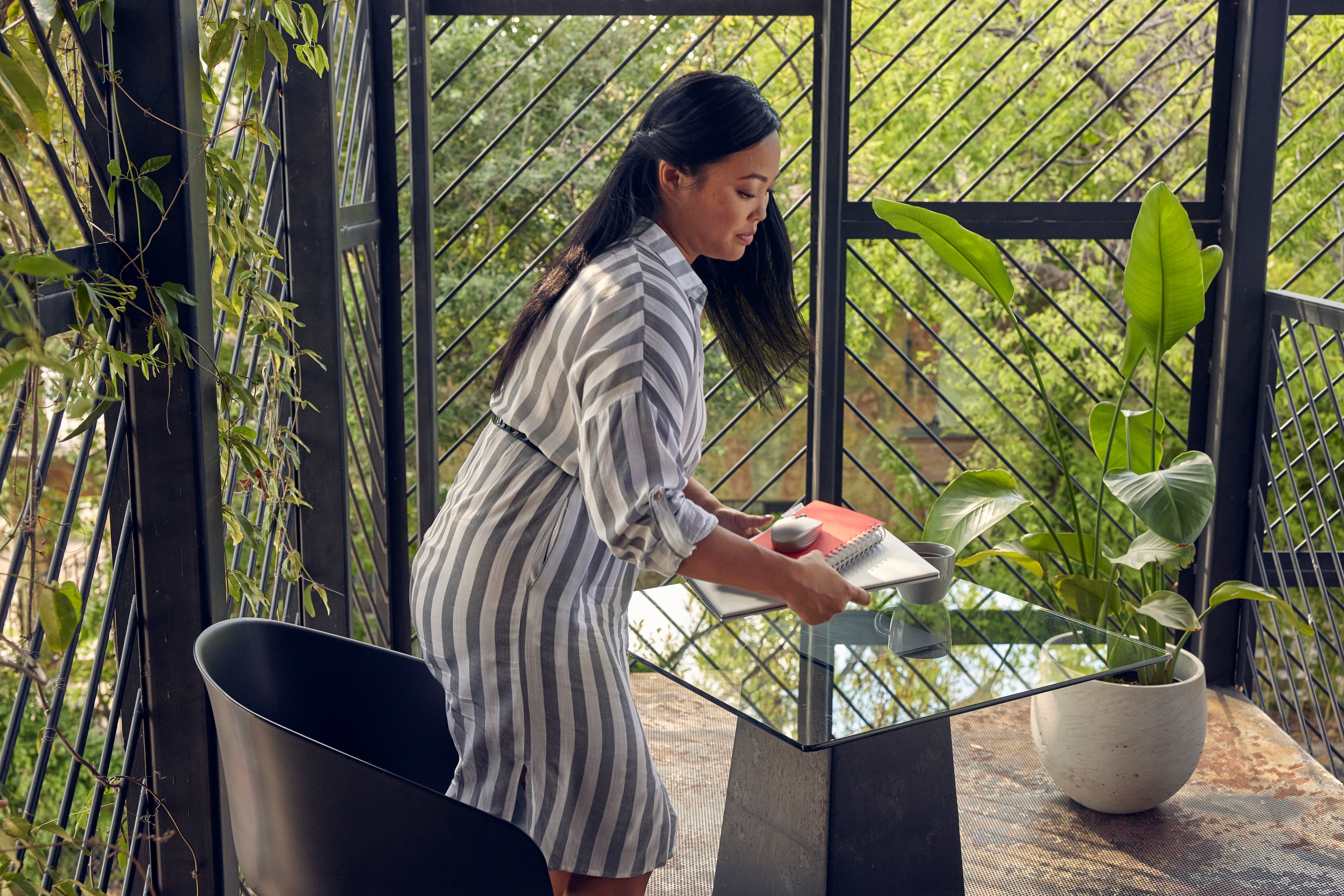 Kobieta w pasiasta sukience, stojąca przy stole. Zielone rośliny na balkonie. Słoneczny dzień.