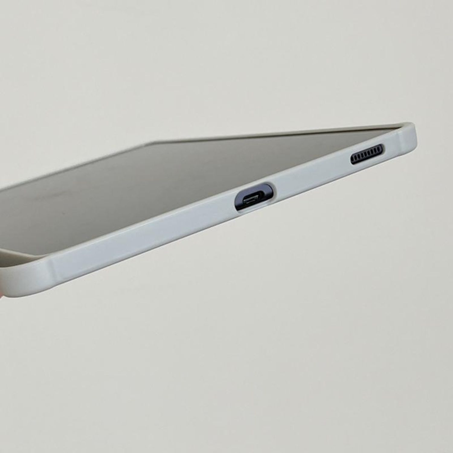 Funda de tableta gris claro. Vista lateral mostrando puertos y botones. Sujetada por una mano.