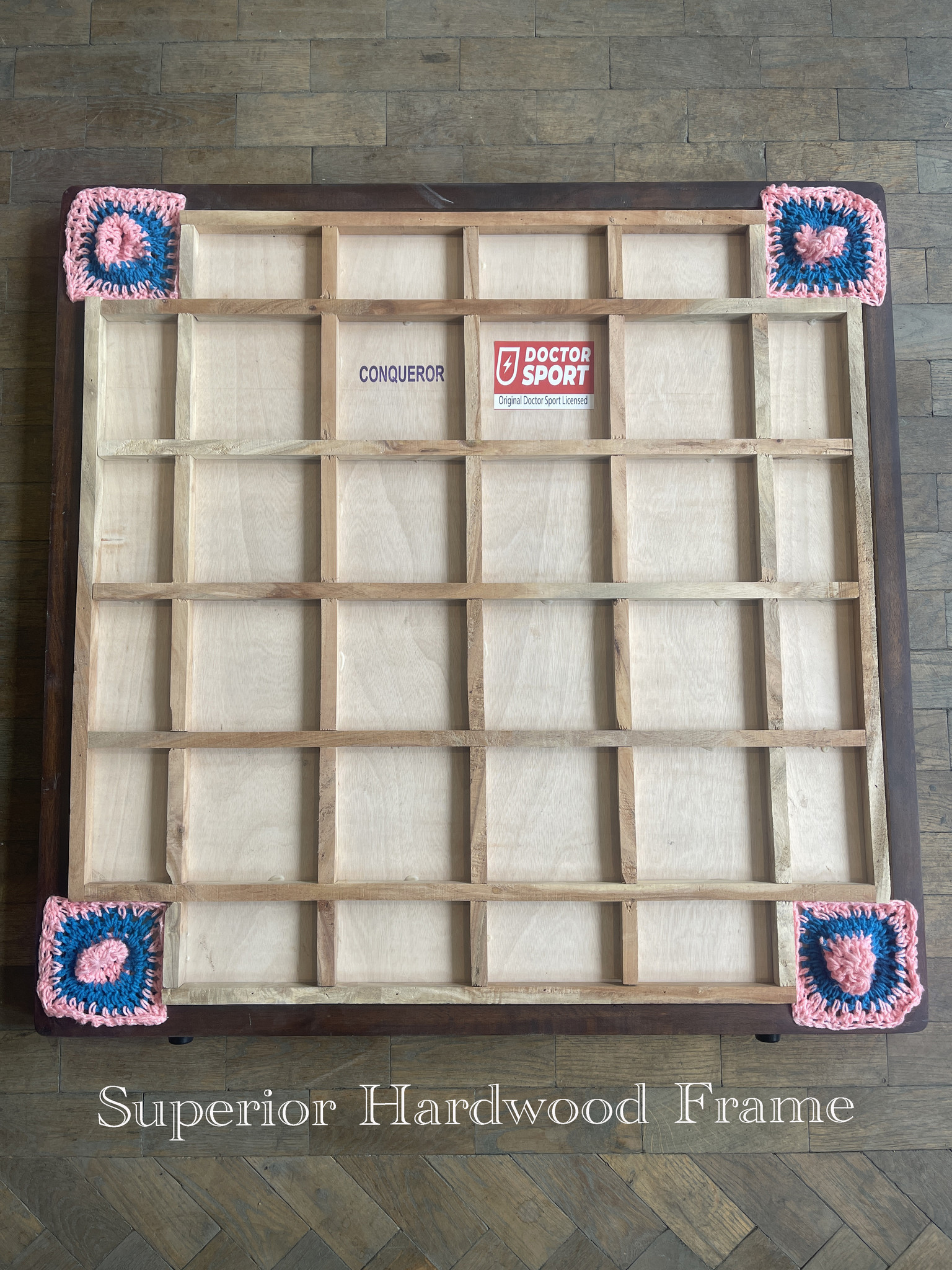 Ein Holzbrettspiel mit rosa und blauen gehäkelten Ecken. Der Text 'CONQUEROR' ist sichtbar.