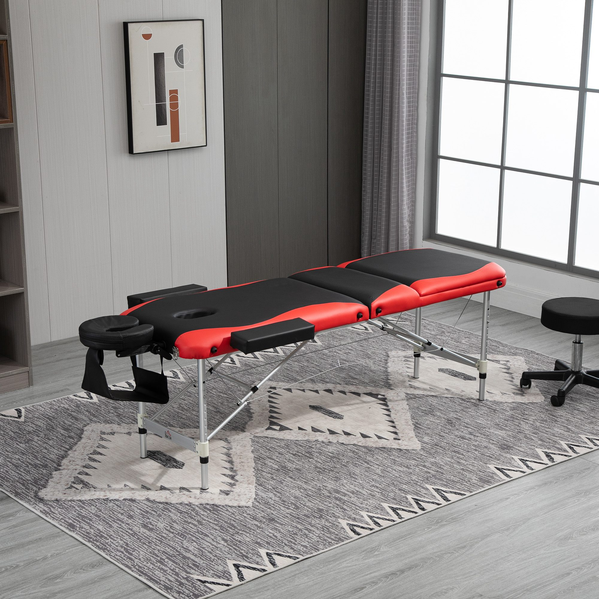 Massagetisch: rot und schwarz, mit Kopfstütze, Armlehnen und einem Hocker. Auf einem Teppich in einem Raum mit Fenster.