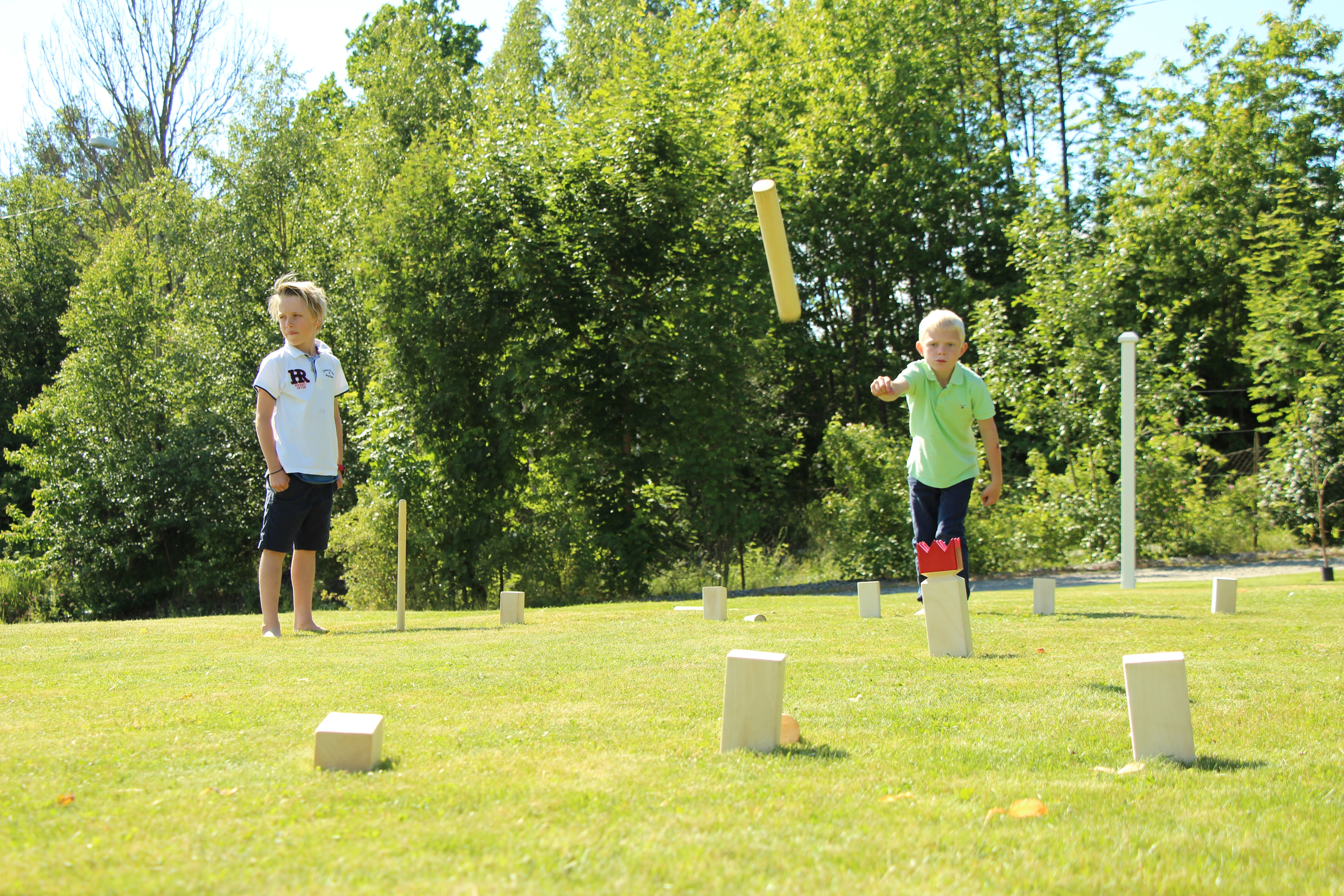 Zwei Jungen spielen ein Holzspiel auf einer grünen Wiese. Einer wirft einen gelben Stock.