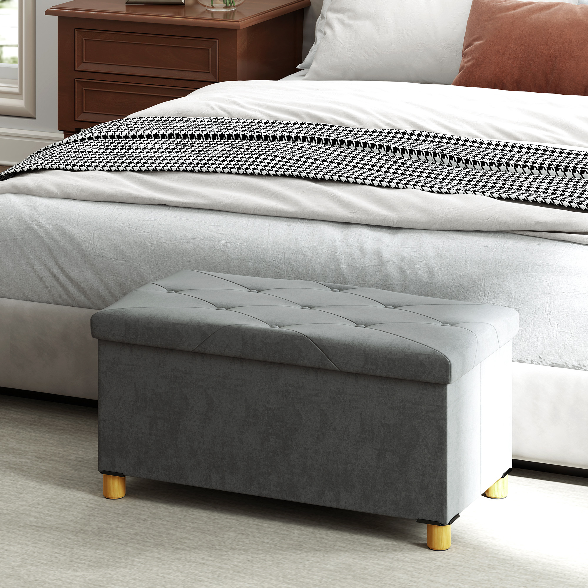 Eine graue Aufbewahrungsottomane mit goldenen Beinen steht vor einem Bett mit Kissen und einer Decke.