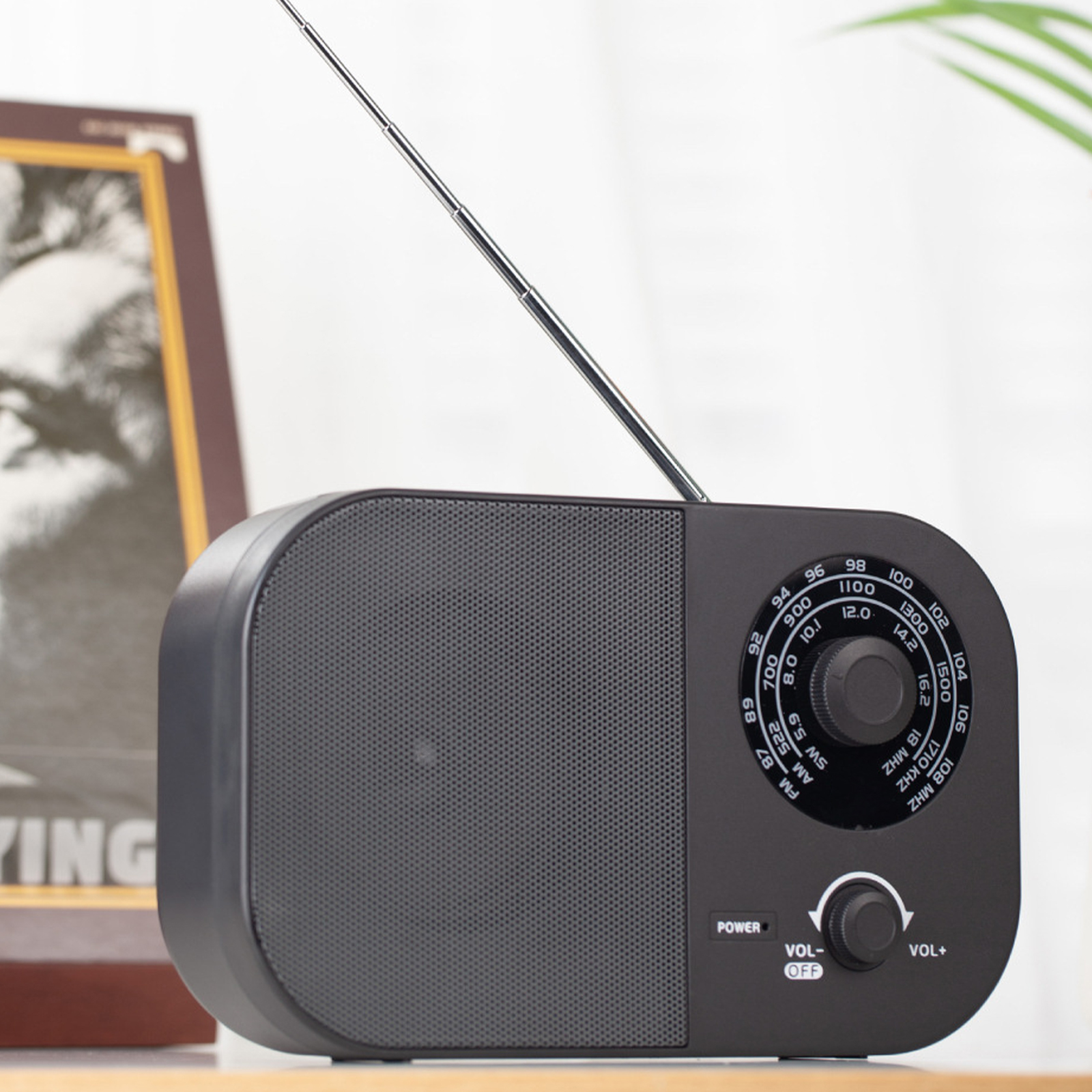 Ein schwarzes Radio mit Antenne und Bedienelementen, das auf einer Holzoberfläche steht. Ein Foto im Hintergrund.