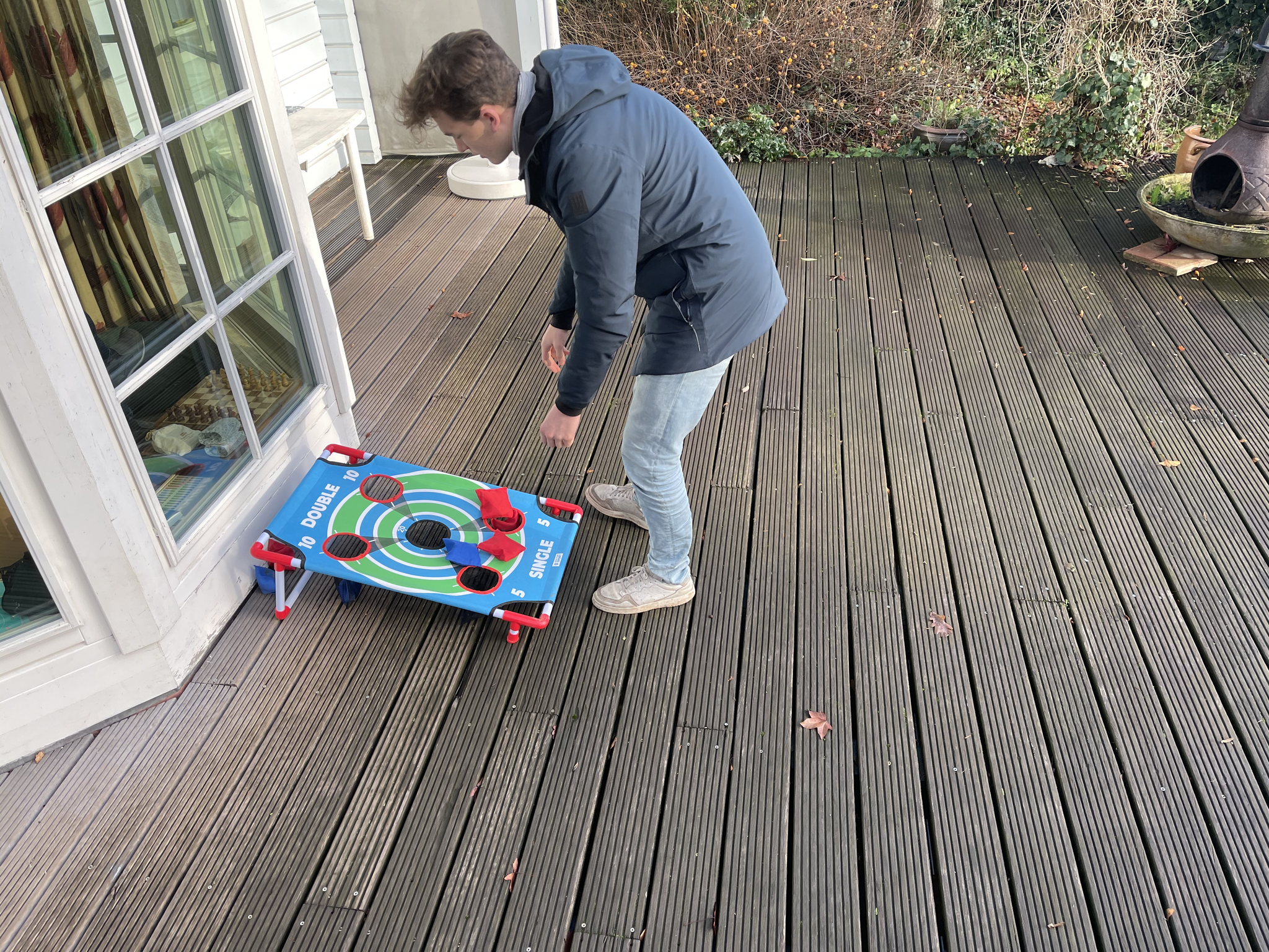 Ein Mann beugt sich über ein Cornhole-Board auf einer Holzterrasse neben einer Glastür.