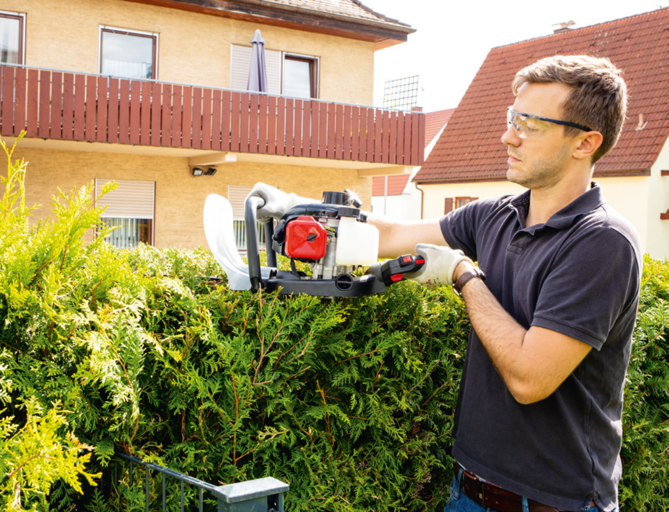 Mann benutzt Heckenschere an grüner Hecke, trägt Schutzbrille und Handschuhe.