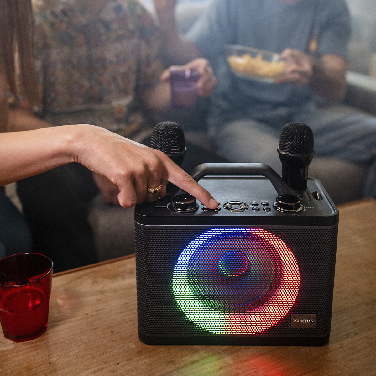 Palec osoby naciska przycisk na głośniku do karaoke. Na stole są ludzie, chipsy i napój.