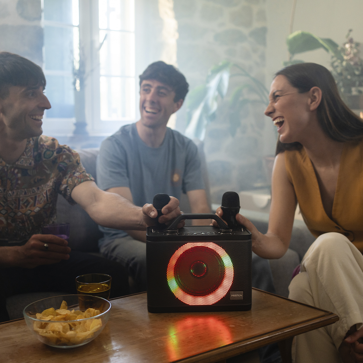 Trzy osoby śmieją się, trzymając mikrofony w pobliżu głośnika z kolorowymi światłami.