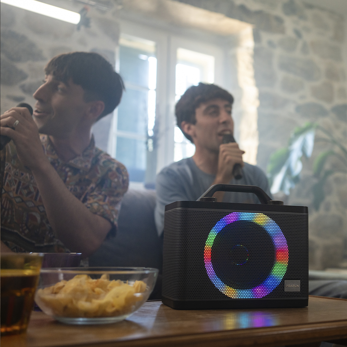 Dwóch mężczyzn z mikrofonami, głośnik z kolorowymi światłami i przekąskami na stole.