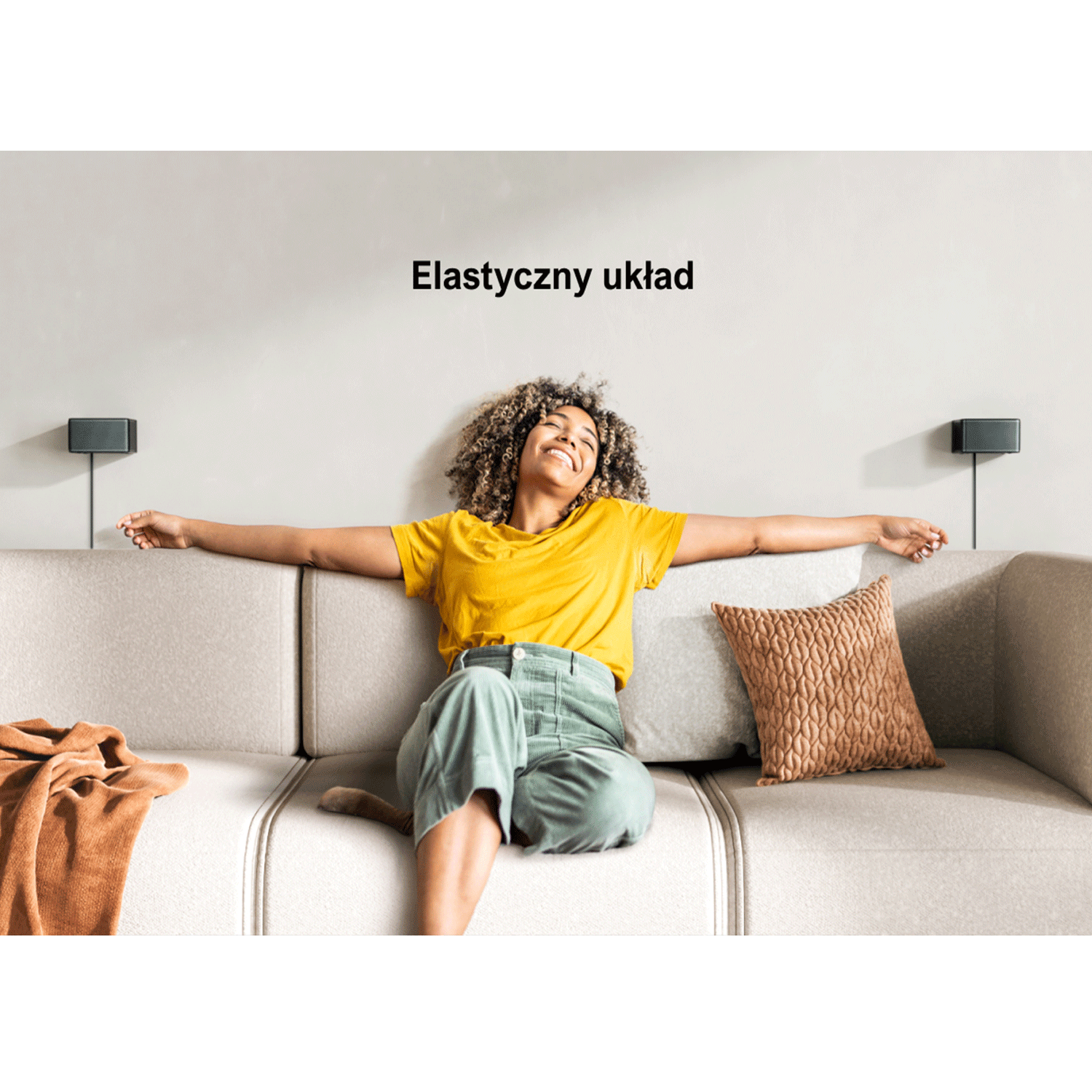 Kobieta na sofie z głośnikami na ścianie. Brązowy koc i poduszka.