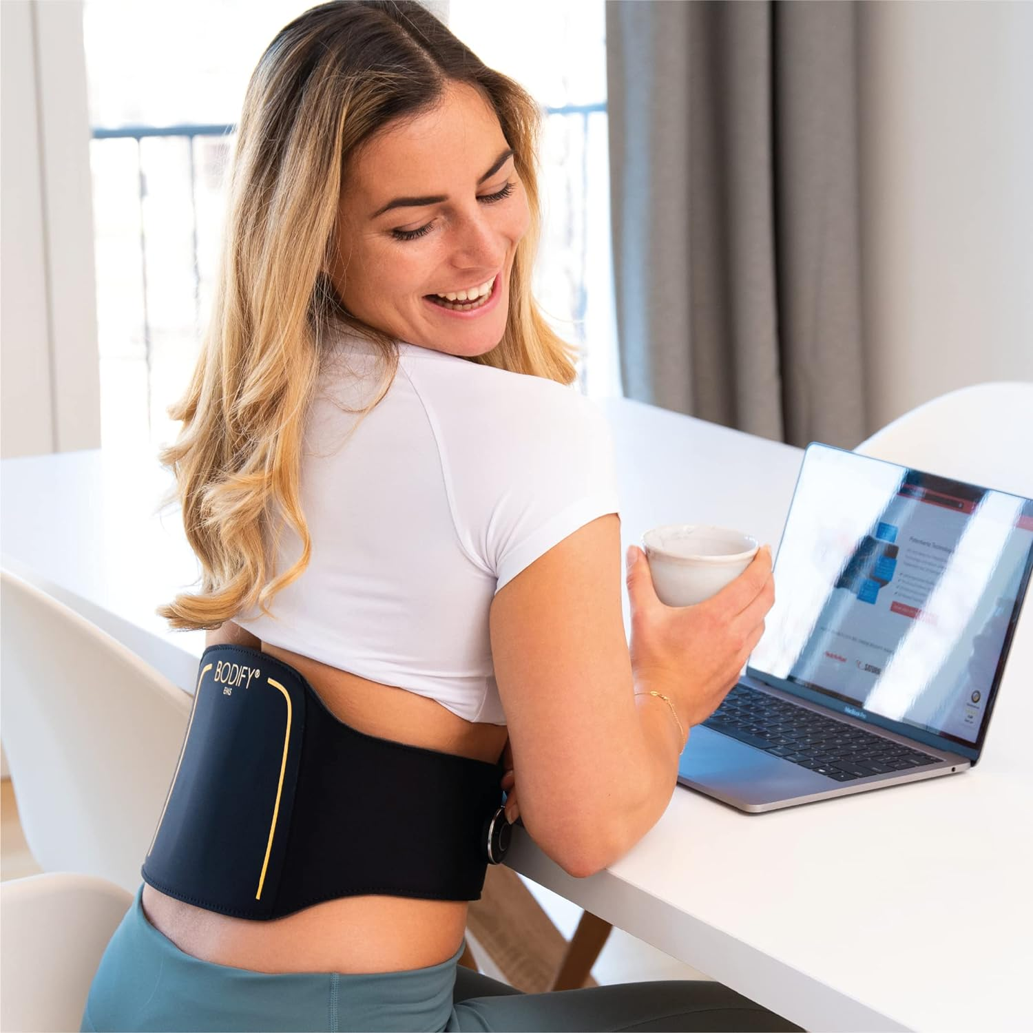Frau mit Rückenstütze, Laptop und Tasse. Hintergrund: Tisch, Vorhänge und Wand.